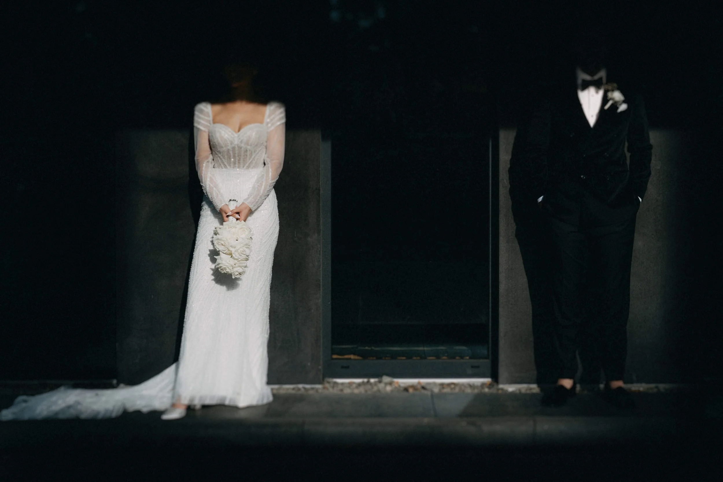 Ein Brautpaar steht mit Abstand nebeneinander, die Braut trägt ein langes weißes Hochzeitskleid mit Spitze, die Angetraute trägt einen schwarzen Anzug mit Fliege, vor ihnen befindet sich ein dunkler Hintergrund.