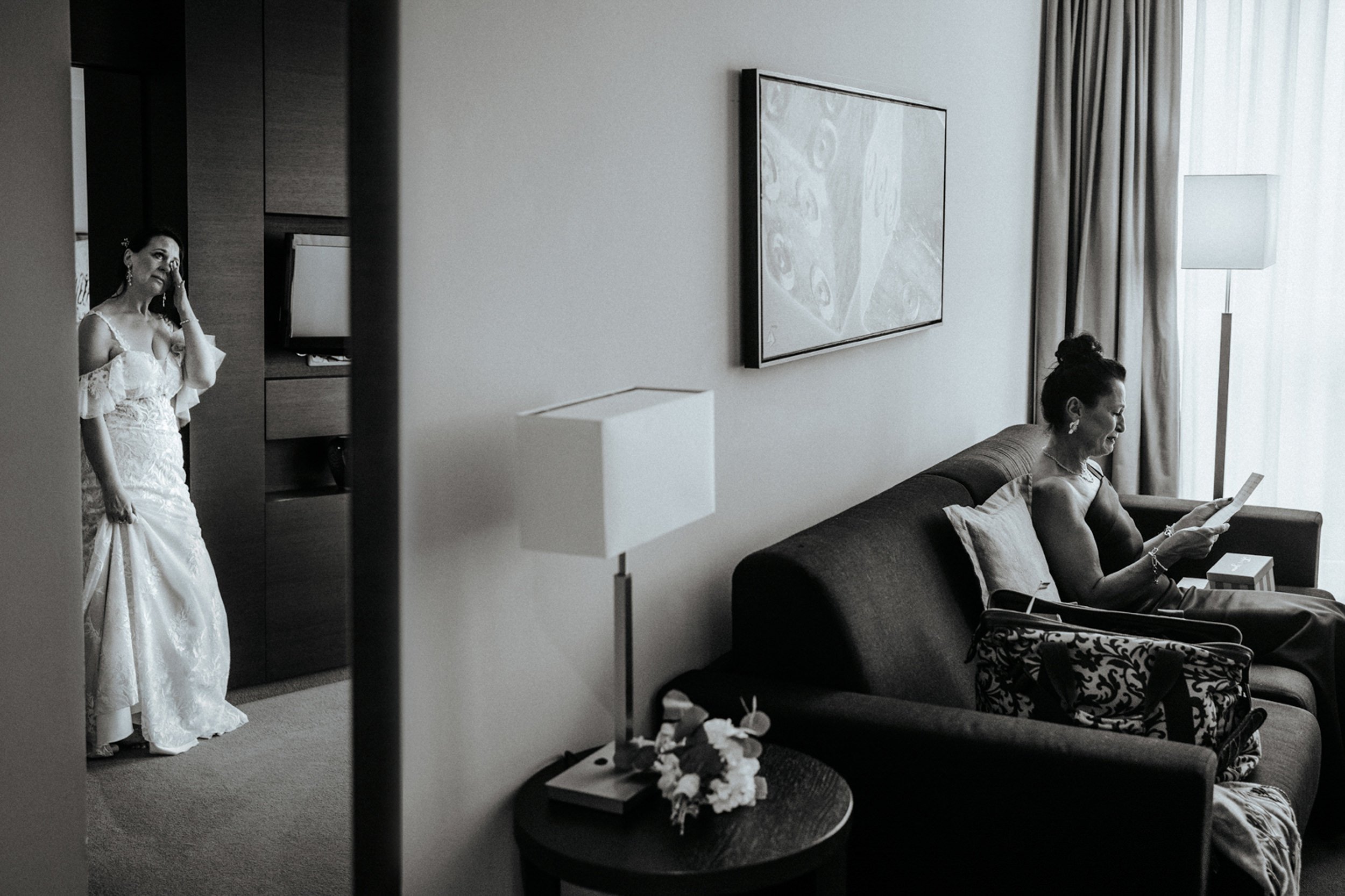 Zwei Frauen in einem Hotelzimmer. Eine Frau steht in einem weißen Hochzeitskleid und ist emotional ergriffen, während die andere Frau im schwarzen Sitz bequem sitzt und ein Buch liest.