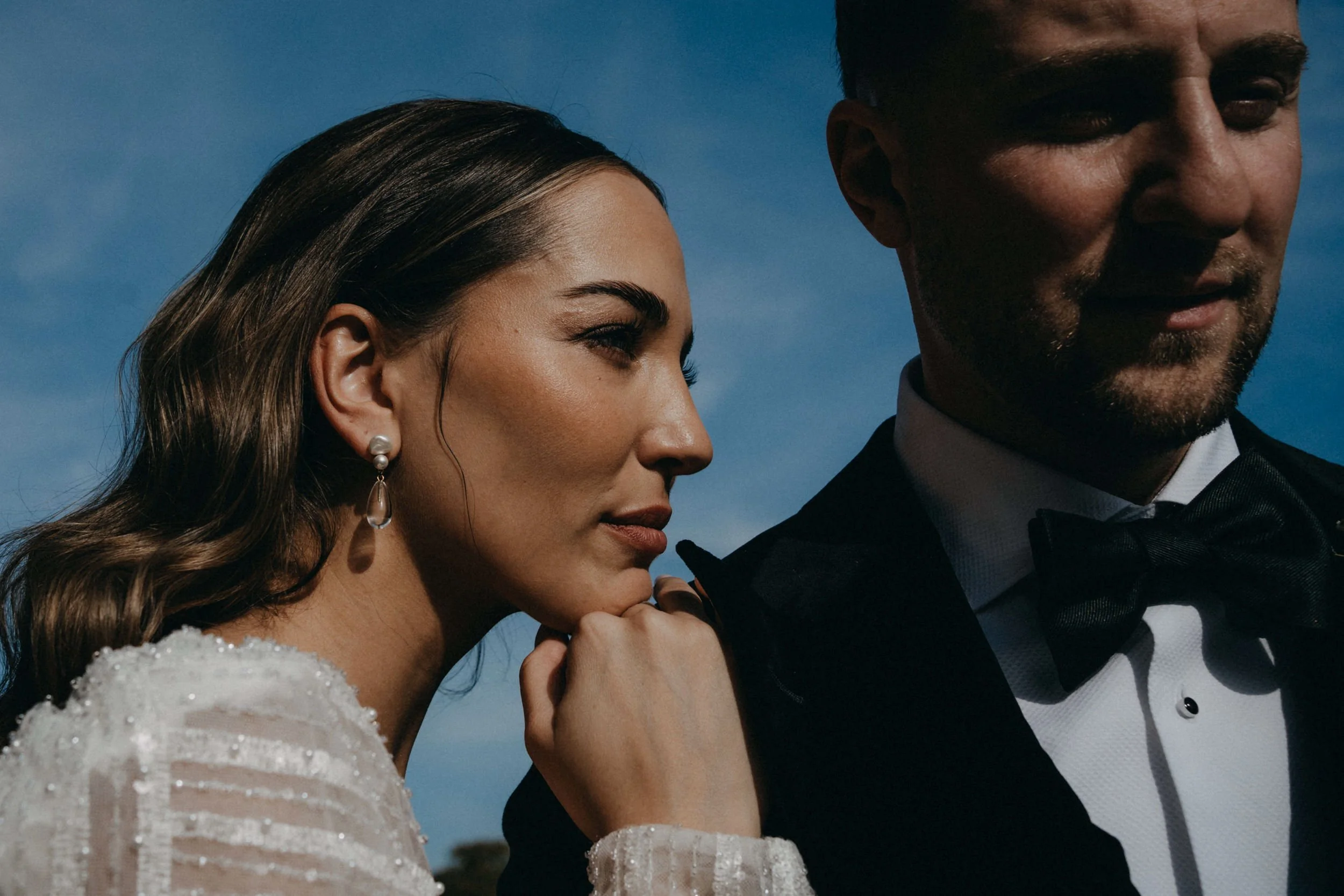 Ein elegantes Paar, eine Frau und ein Mann, stehen eng beieinander im Freien mit blauem Himmel im Hintergrund. Die Frau trägt Perlenohrringe und eine weiße, elegante Kleidung, während der Mann einen schwarzen Smoking mit Fliege trägt. Sie scheinen ei
