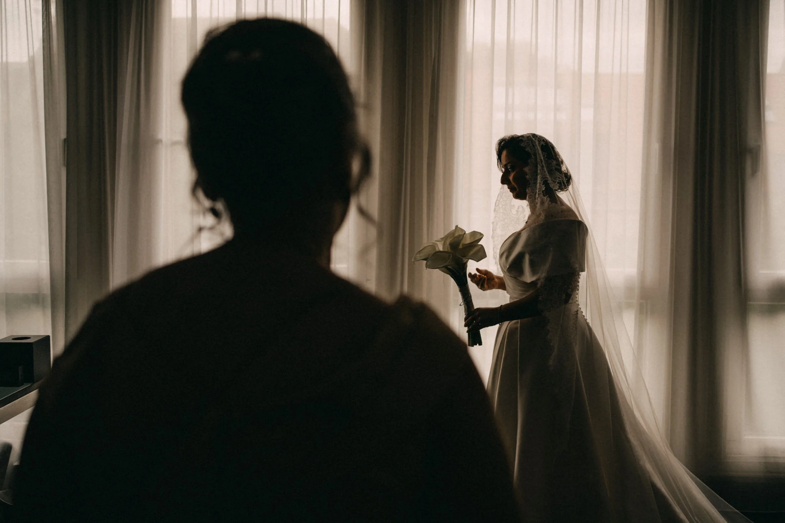 Eine Hochzeitsszene mit einer Braut, die in einem Raum mit hohen Fenstern und weißen Vorhängen steht. Die Braut trägt ein weißes Kleid und einen Schleier, hält einen Blumenstrauß in der Hand, während eine männliche Person im Vordergrund sitzt.