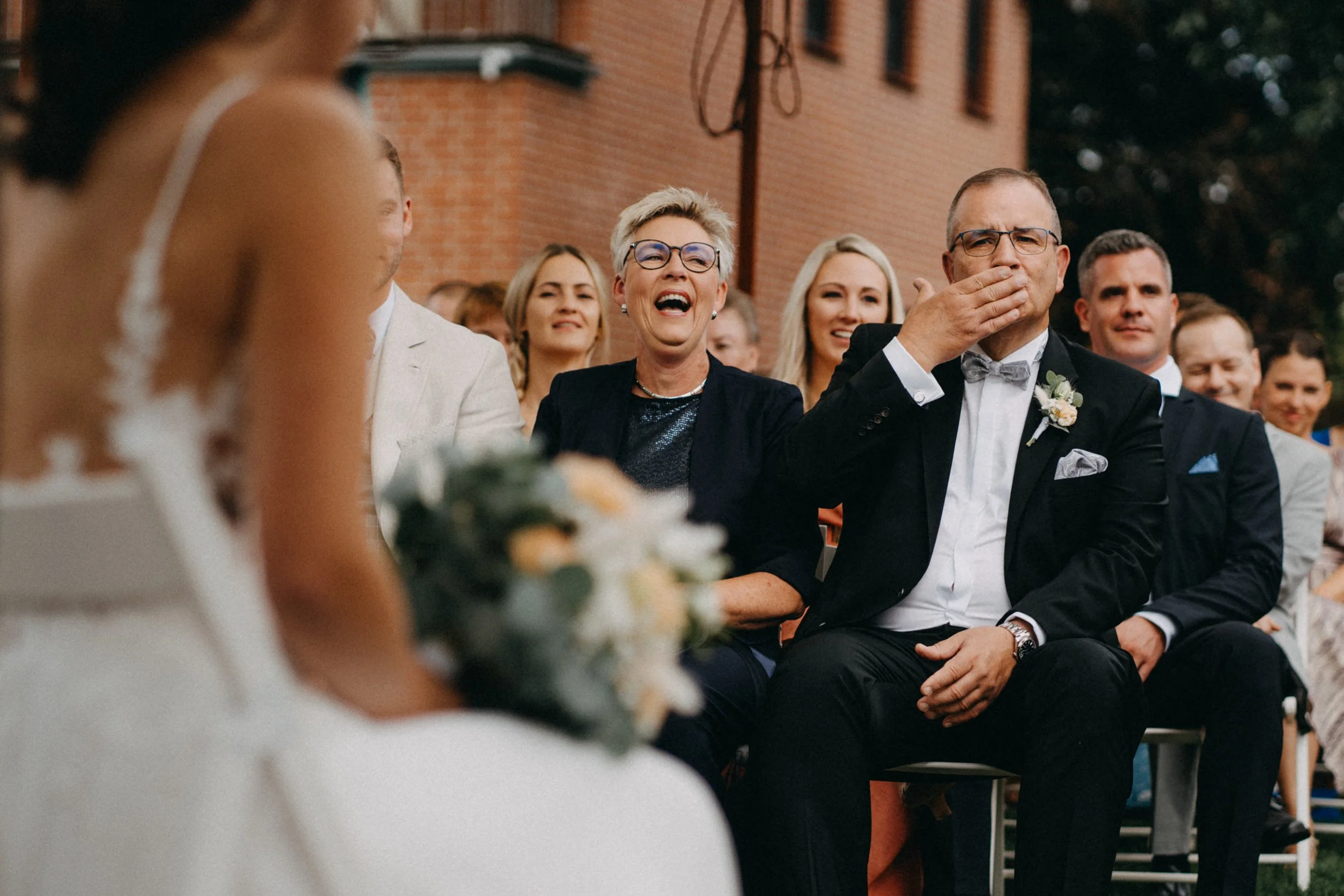 Eine Gruppe von Menschen bei einer Hochzeit, mit einem Mann in Smoking und einer lachenden Frau im Mittelpunkt.
