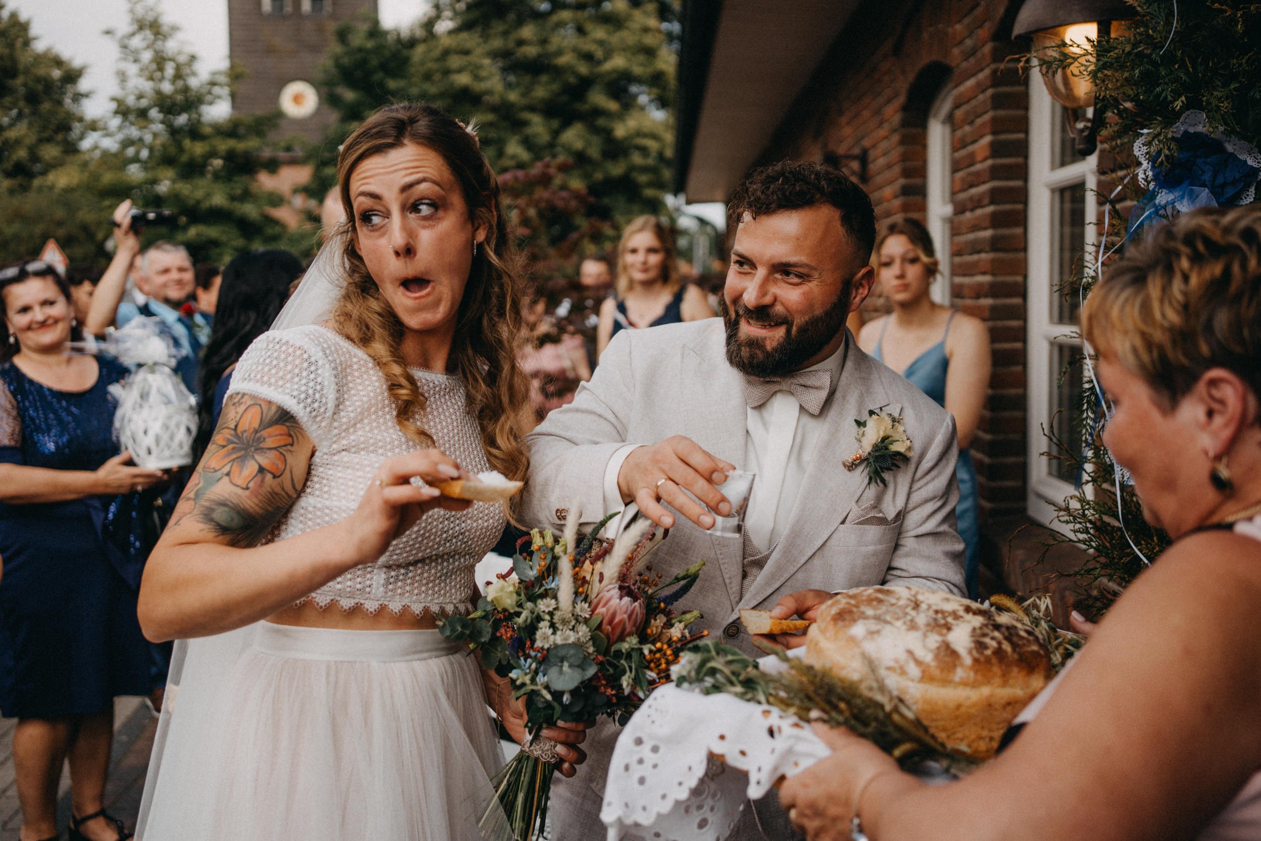 Hochzeitspaar mit Gästen beim Essen im Freien, festliche Atmosphäre, Gäste in Festkleidung, blühende Pflanzen, Ziegelhäuser im Hintergrund.