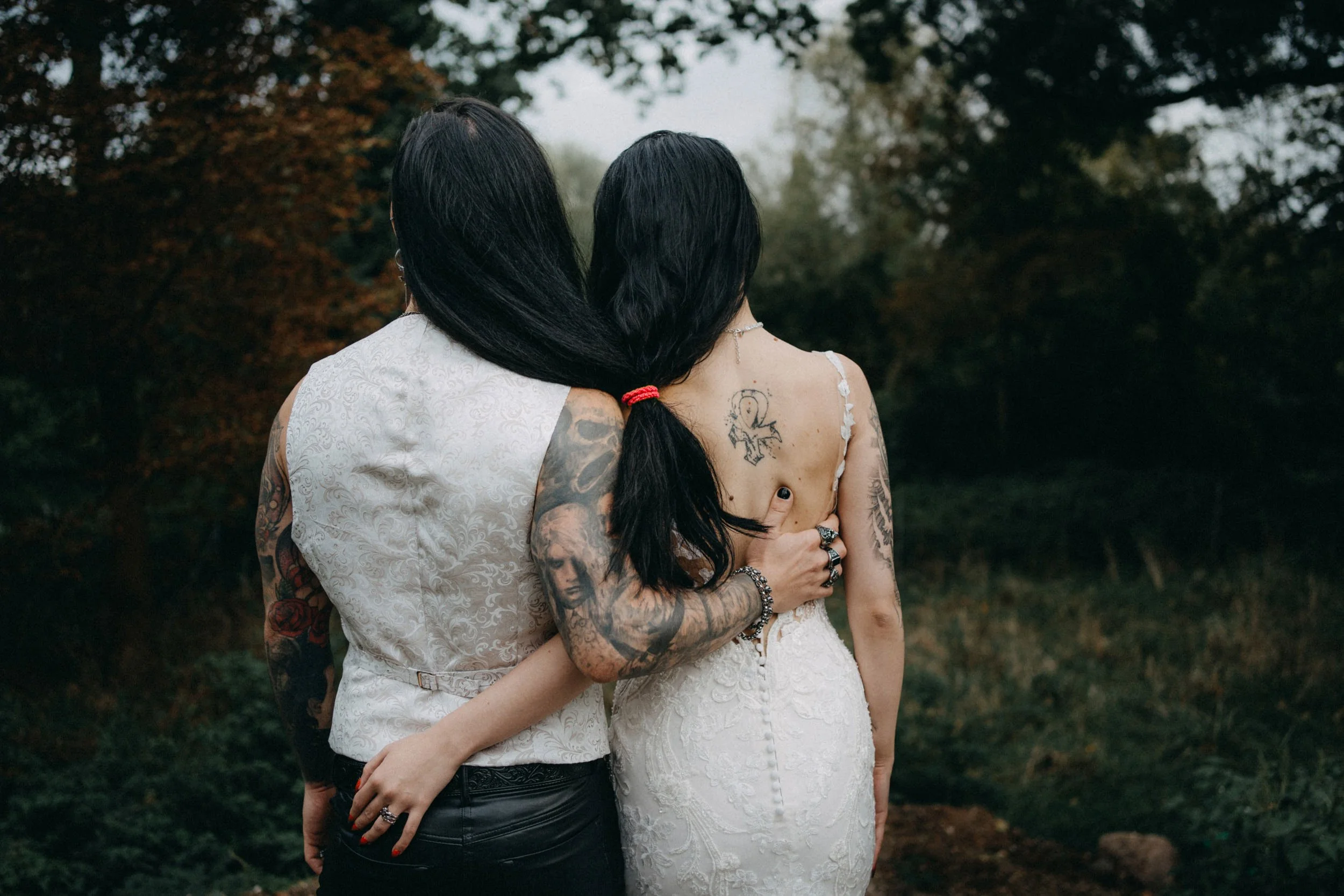 Zwei Frauen mit Tattoos umarmen sich im Freien vor einem herbstlichen Baum.
