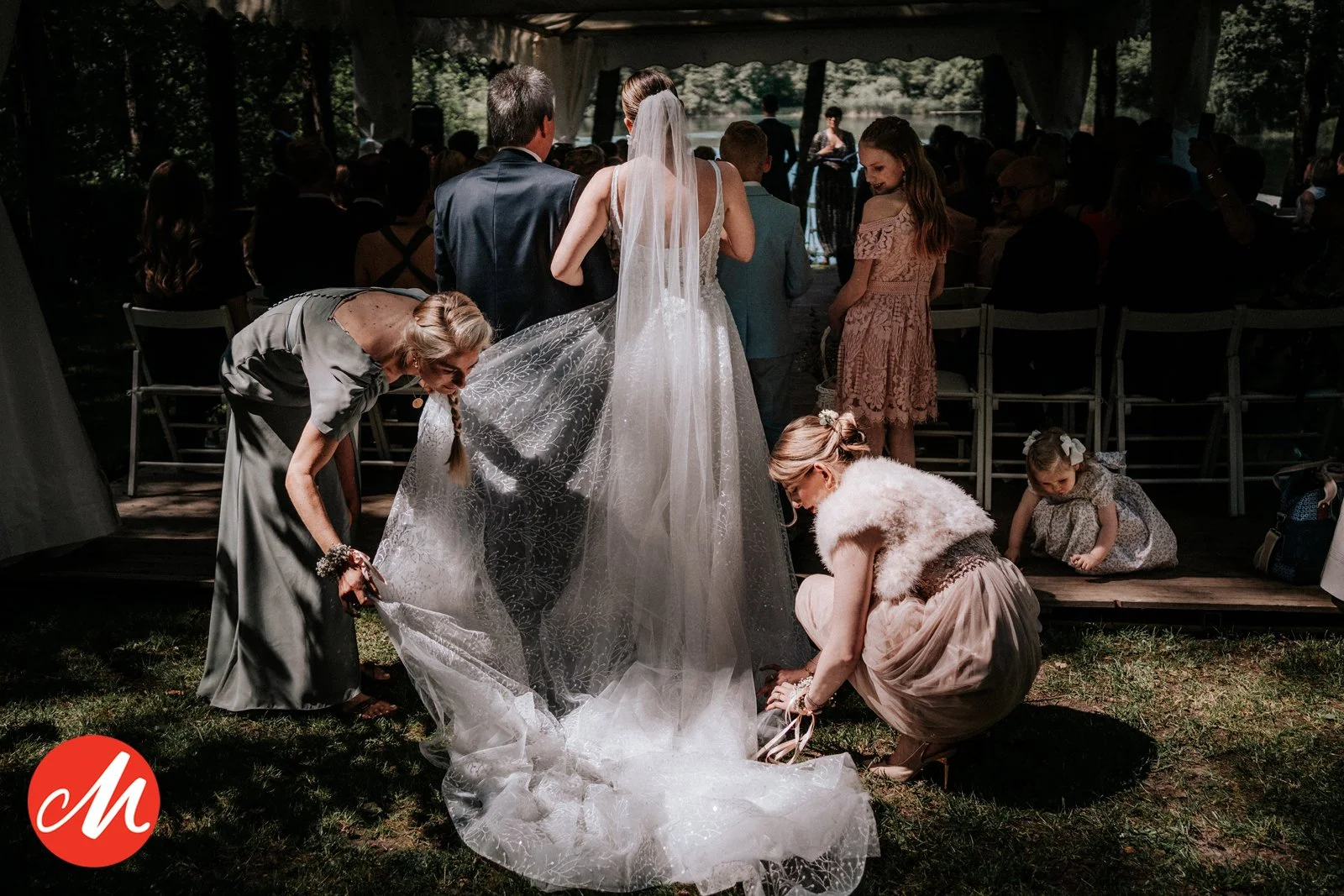 Menschen bei einer Hochzeit, die den Brautkleid voller Detail inspizieren, außerhalb unter einem Pavillon, mit vielen Gästen im Hintergrund