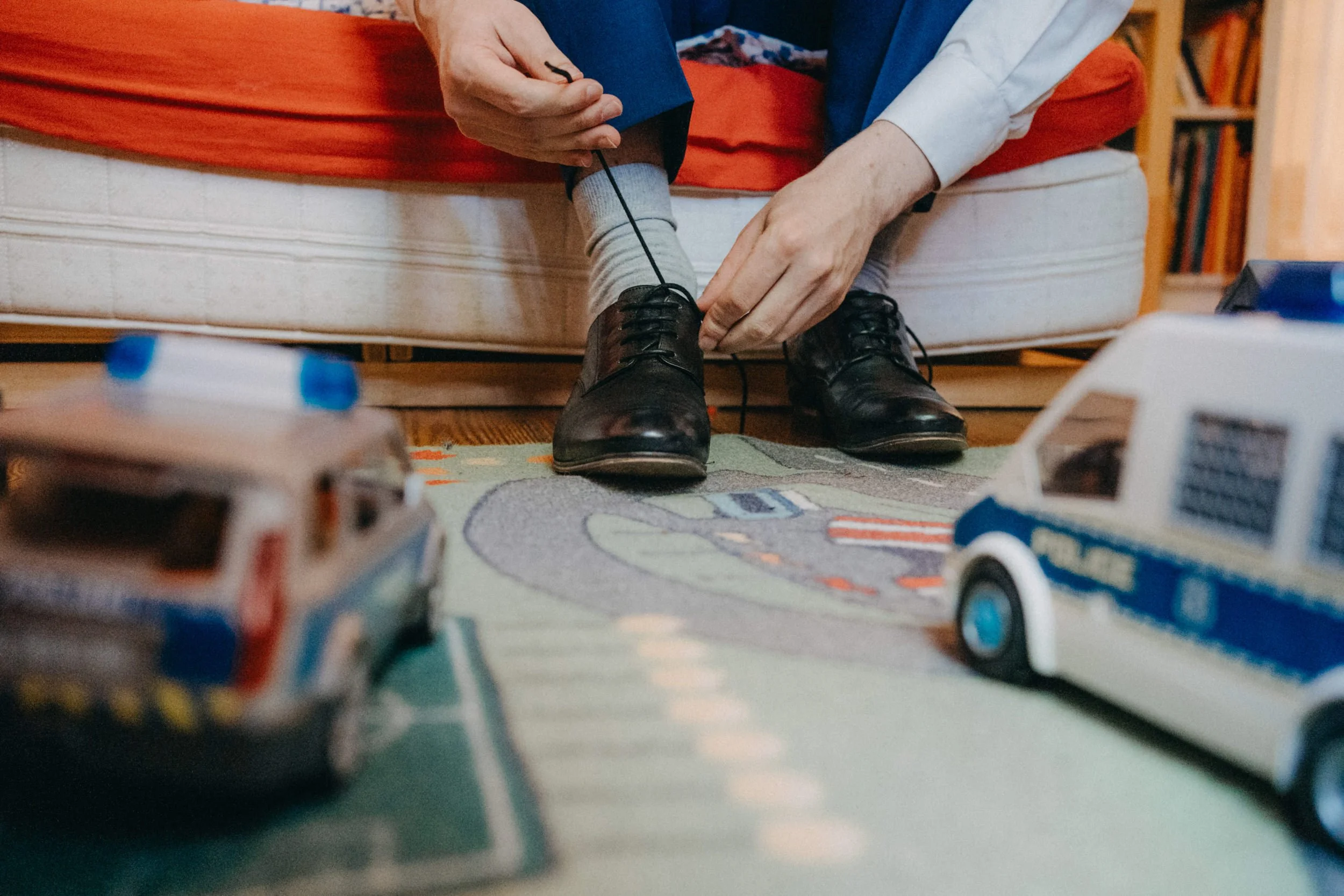 Person, die Schuhe bindet, auf einem roten Sofa sitzend, im Hintergrund Bücherregale, Vordergrund mit Spielzeugautos.