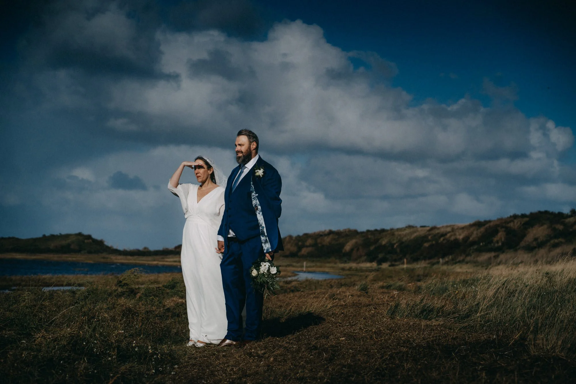 Ein Paar bei einer Hochzeit, das im Freien in einer ländlichen Küstenregion steht, die Frau in einem weißen Kleid, der Mann in einem blauen Anzug, während sie auf die Ferne blickt.
