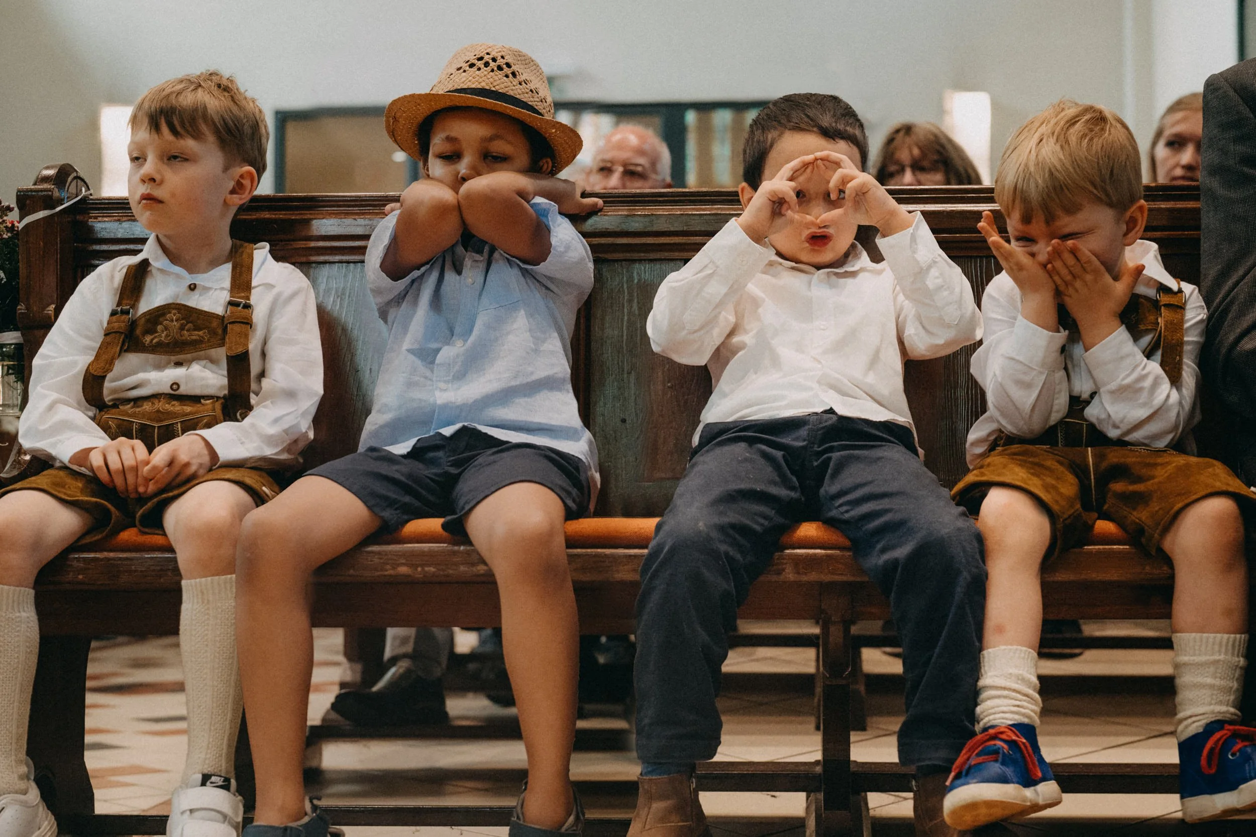 Vier Kinder sitzen auf einer Bank, die Kinder sind in traditionellen Trachten gekleidet. Die Kinder auf der linken Seite sieht man nur halb. Die drei rechten Kinder machen verschiedene Gesichter und Gesten, während zwei andere Erwachsene im Hintergru