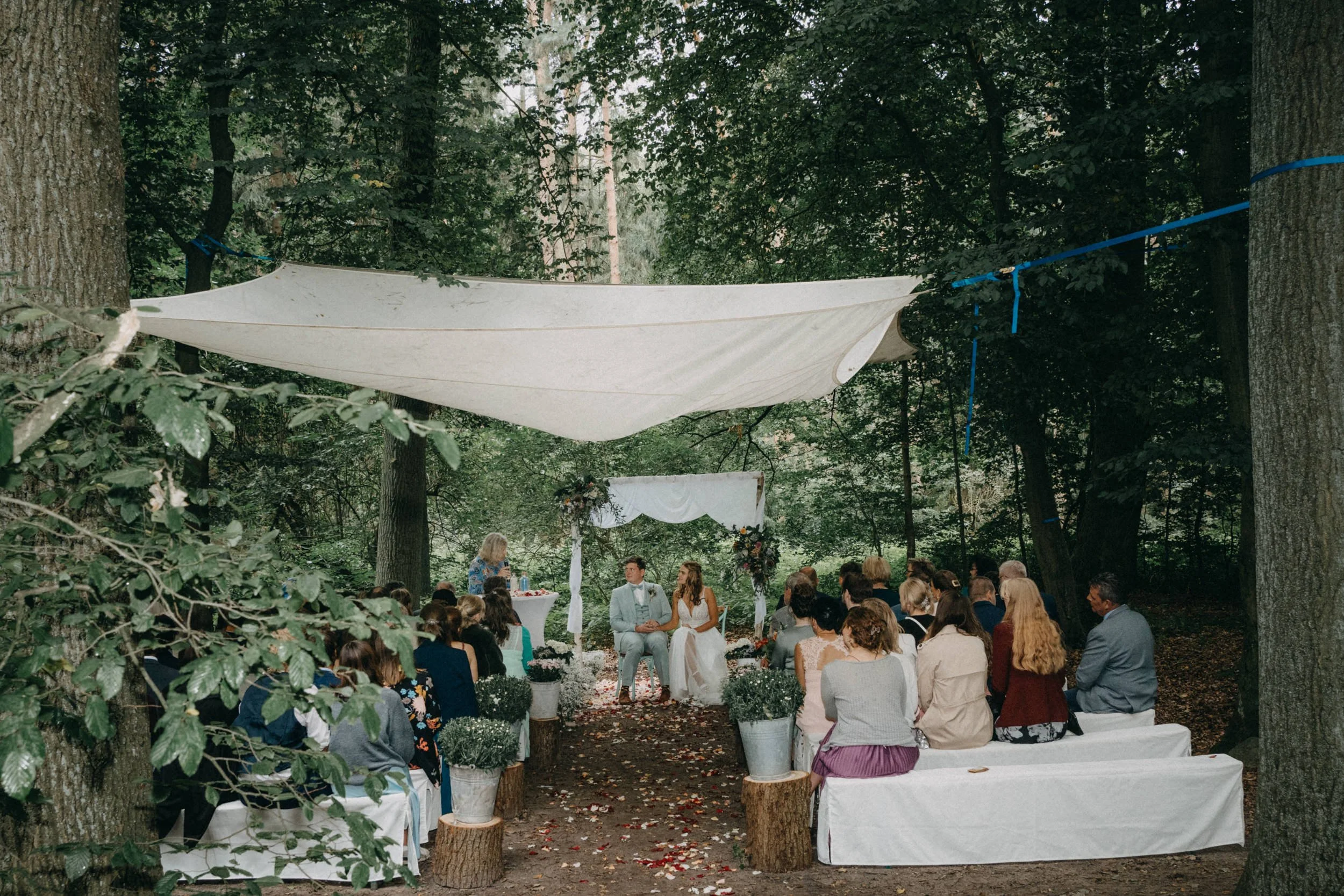 Eine Zeremonie im Freien im Wald mit vielen Gästen, einem Brautpaar, einem Altar und Dekorationen unter einem weißen Sonnen- oder Regenschutz