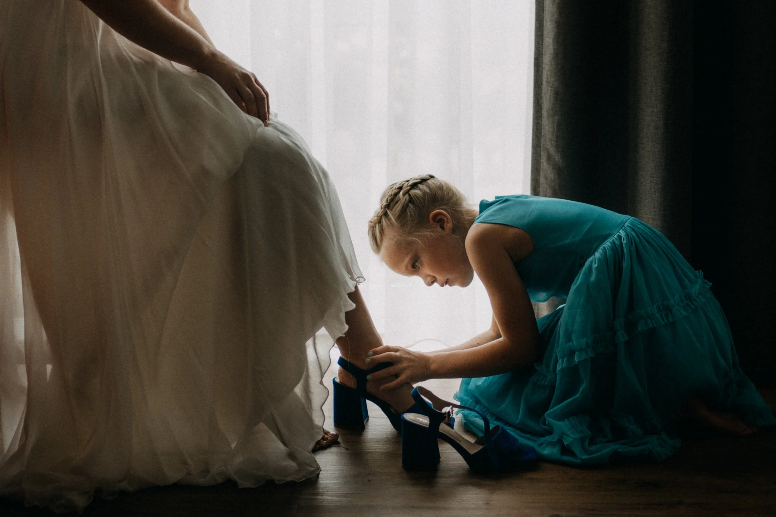 Ein Mädchen in einem blauen Kleid liegt auf dem Boden und bringt eine Kamera in Position, während eine Person im weißen Kleid neben ihr kniet, vermutlich beim Fotografieren oder Filmen.