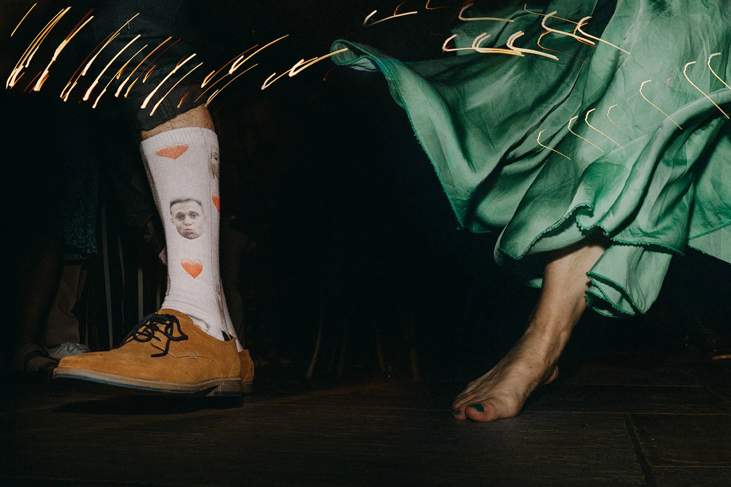Nahaufnahme eines Menschen mit grüner Tüllrock, Orange Schuhe, weißer Socke mit Herzmuster und einem Gesicht aufgenäht, der auf einem hölzernen Boden kniet. Im Hintergrund sind Lichtspuren sichtbar.
