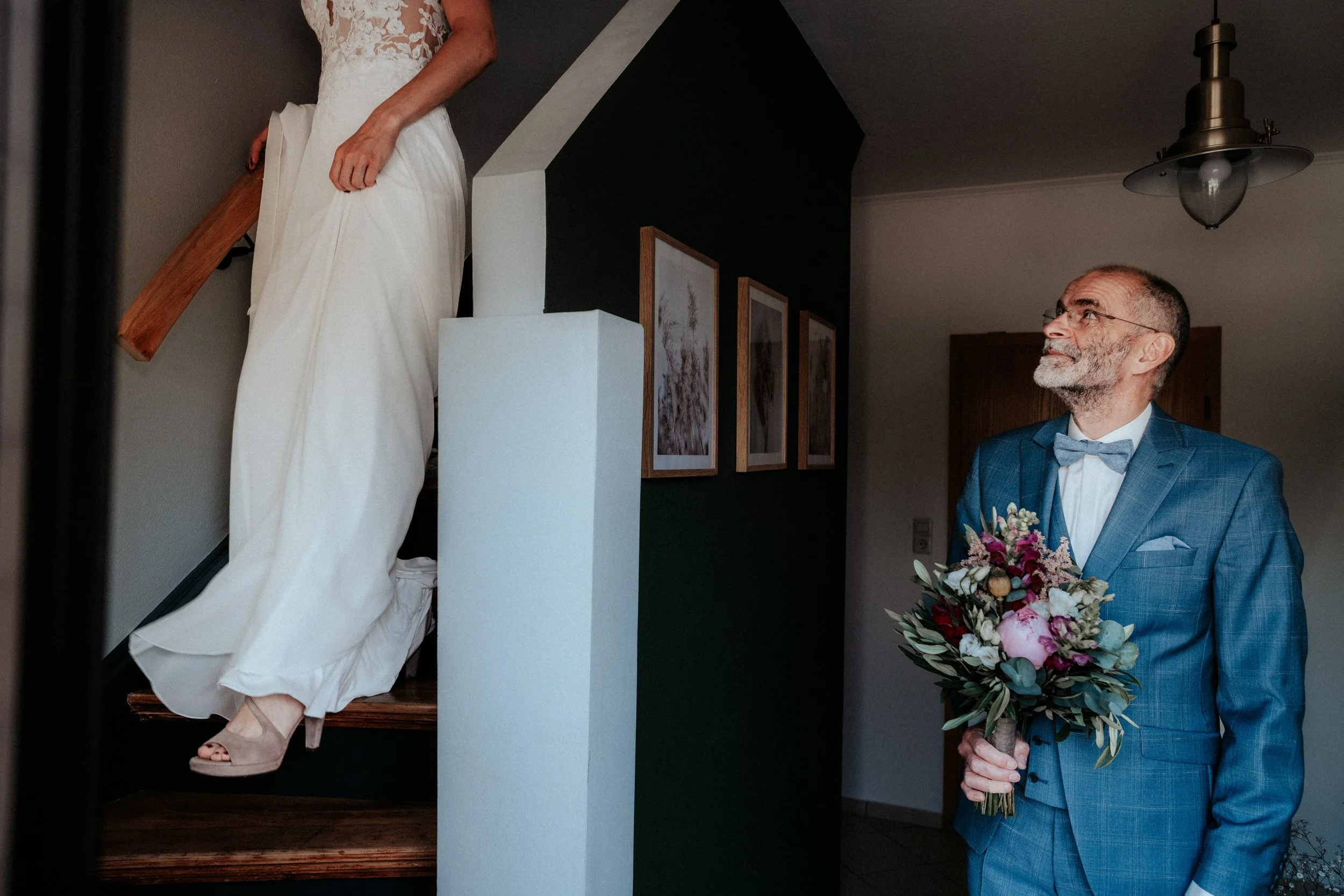 Ein älterer Mann in einem blauen Anzug mit Schleife hält einen Blumenstrauß und schaut zu einer jungen Frau in einem weißen Kleid, die die Treppe hochsteigt.