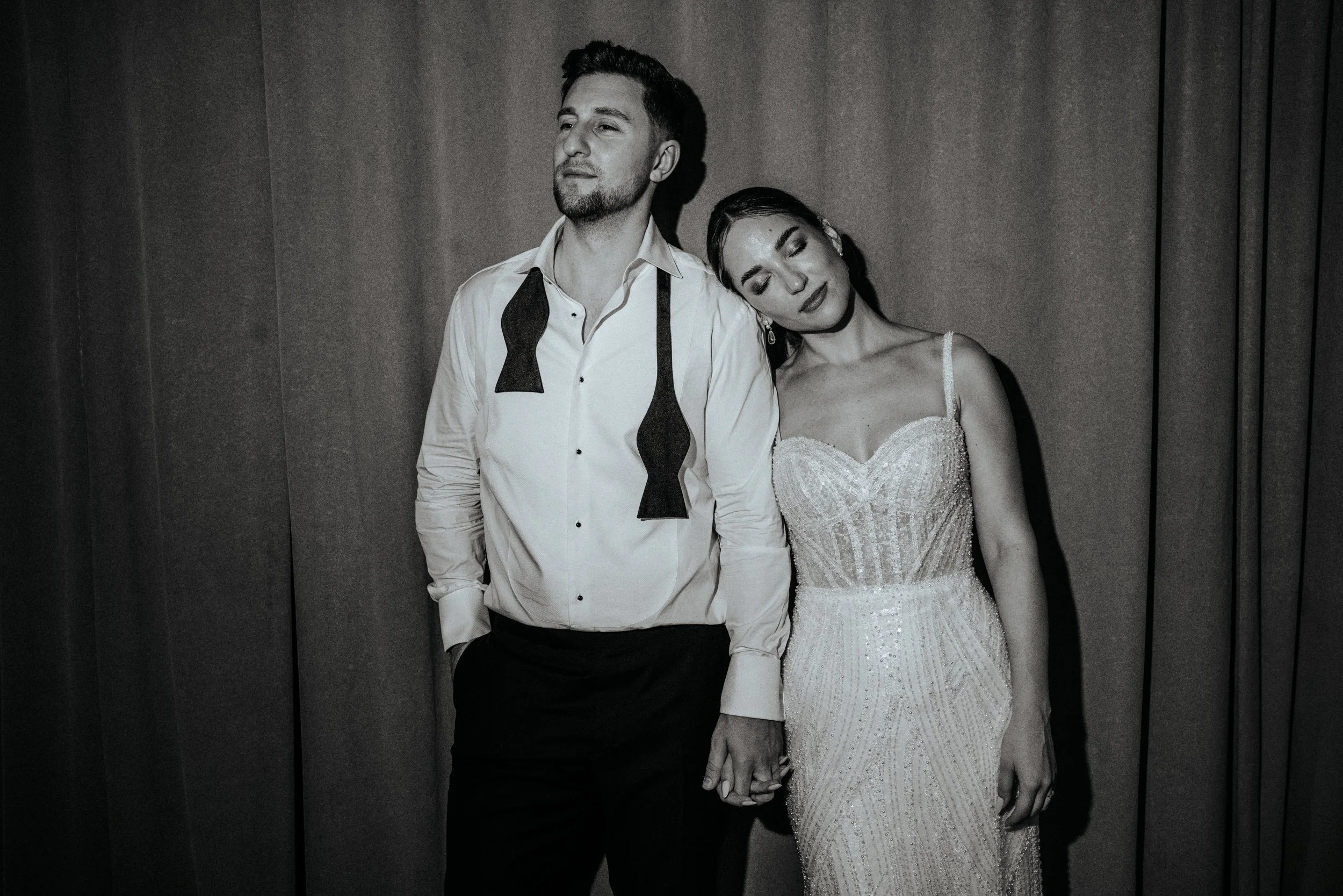 Ein Mann und eine Frau halten Händchen, der Mann trägt ein weißes Hemd mit einer schwarzen Fliege, die um den Hals hängt, die Frau trägt ein elegantes, funkelndes Abendkleid.