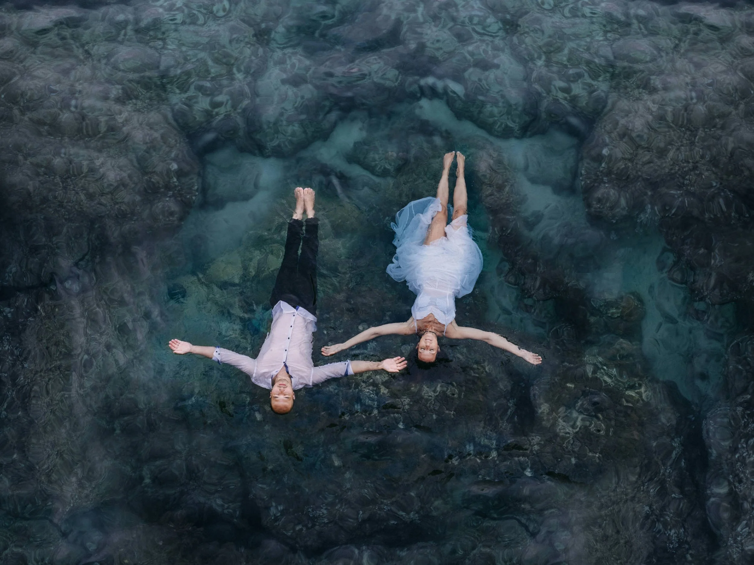 Zwei Menschen liegen auf einem Felsen im Wasser, einer trägt einen weißen Rock, beide haben die Arme ausgestreckt und scheinen entspannt zu sein.
