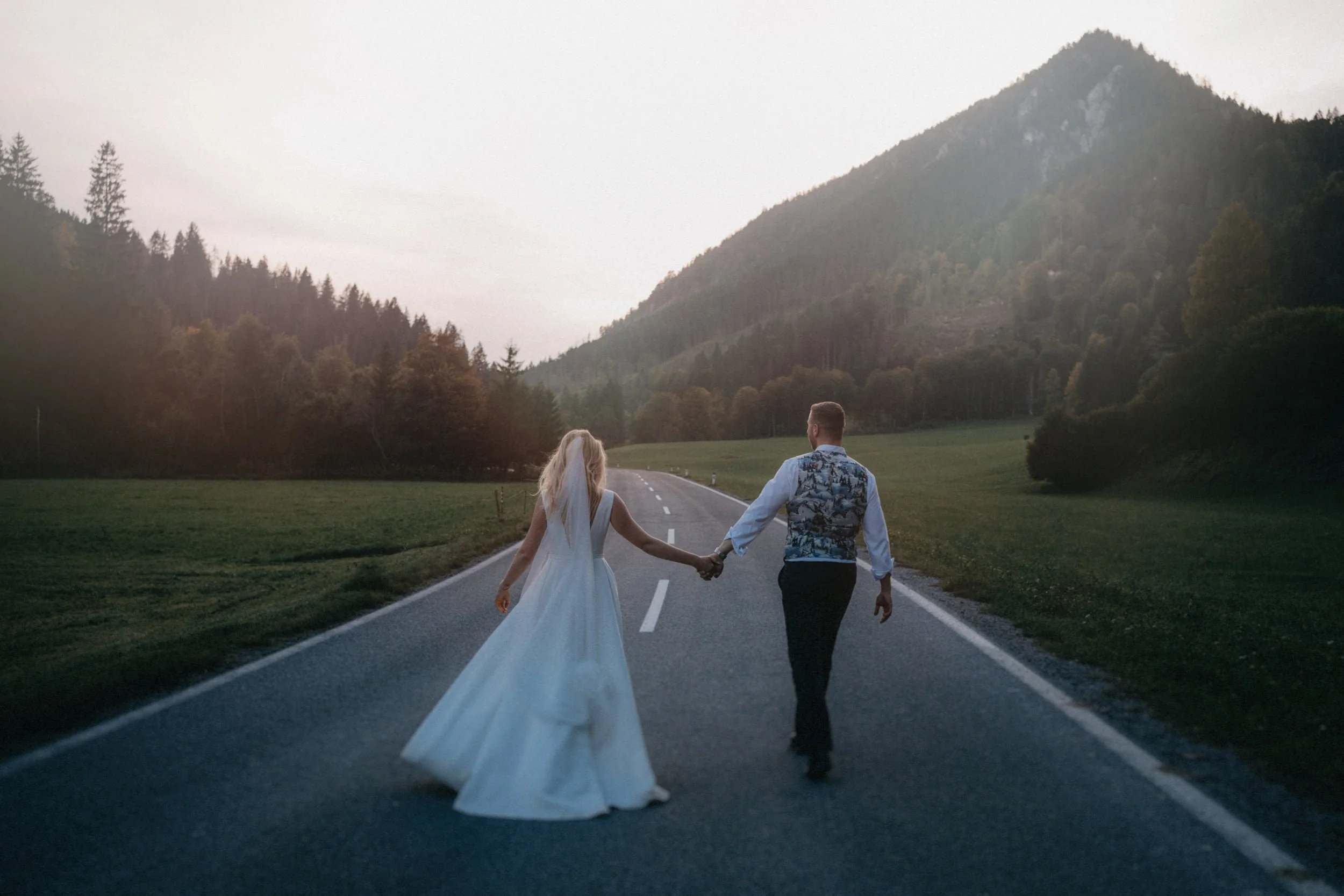 Ein verliebtes Paar, eine Frau in einem Hochzeitskleid und ein Mann, gehen auf einer Landstraße inmitten von Bergen und Wäldern. Sie halten sich an den Händen und schauen sich an.