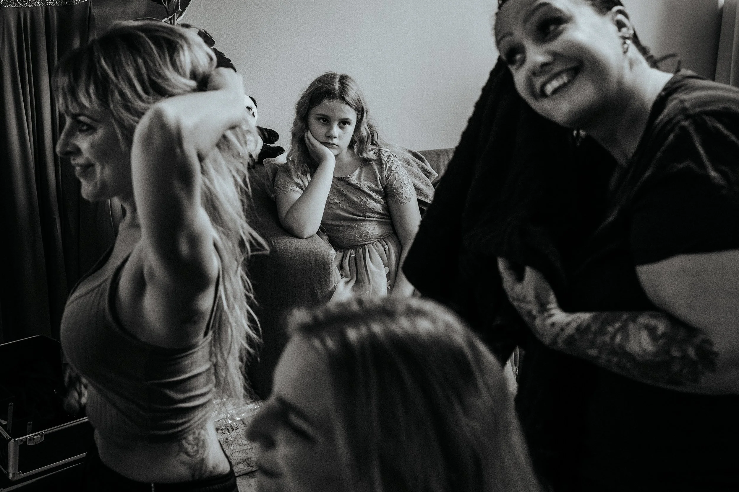 Drei Frauen und ein junges Mädchen stehen in einem Raum. Zwei Frauen lachen, während die dritte Frau und das Mädchen ernst schauen. Die Szene ist in Schwarzweiß gehalten.
