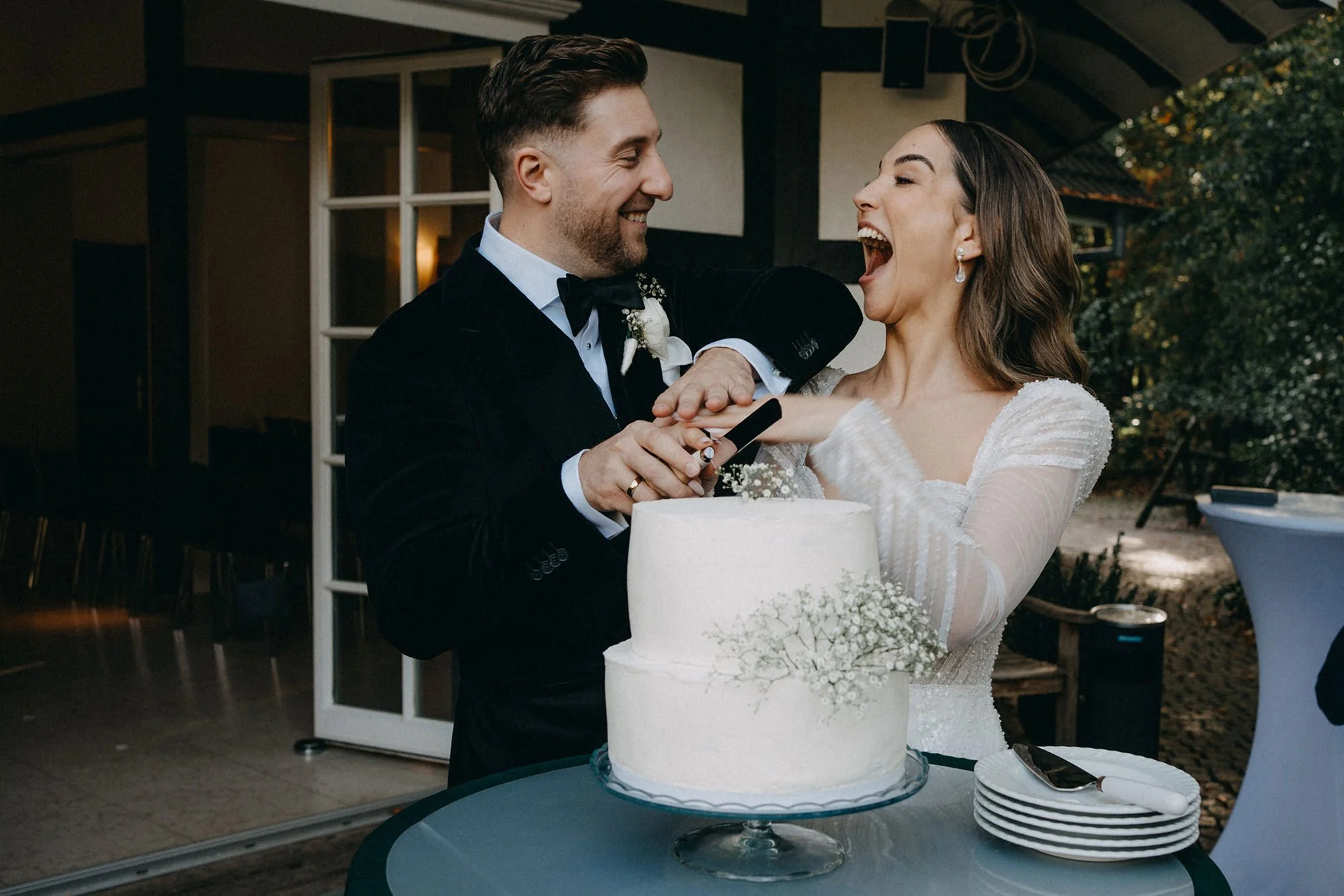 Ein Mann und eine Frau bei einer Hochzeit, lächelnd und lachend, während der Mann dem Brautpaar ein Hochzeitsgeschenk überreicht. Sie stehen vor einem weißen Hochzeitstorten auf einem Tisch, im Hintergrund eine Hütte und Bäume.