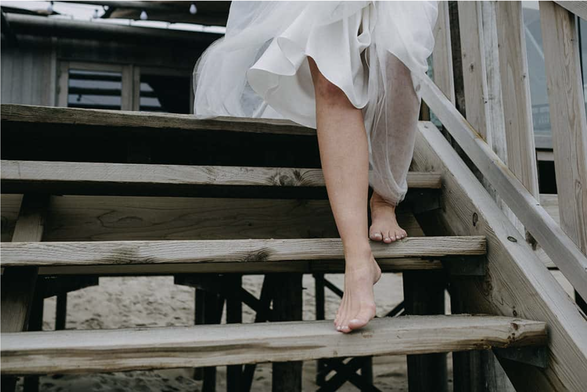 Nahaufnahme eines nackten Beins und Fußes, die auf Treppen aus Holz gehen, mit einem weißen Kleid, das teilweise sichtbar ist.