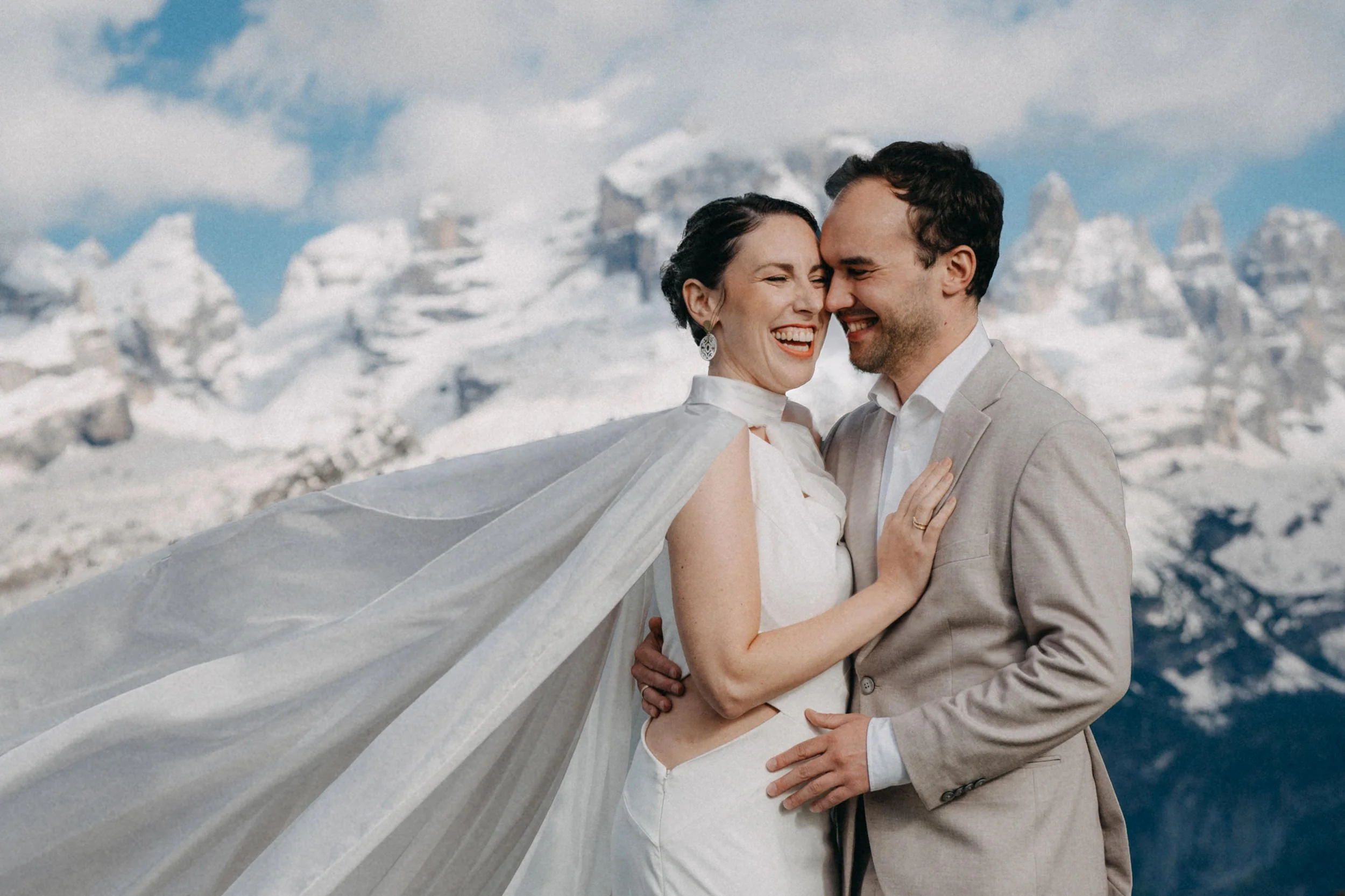 Ein glückliches Paar in Hochzeitskleidung, umarmt sich vor einer schneebedeckten Bergkulisse.