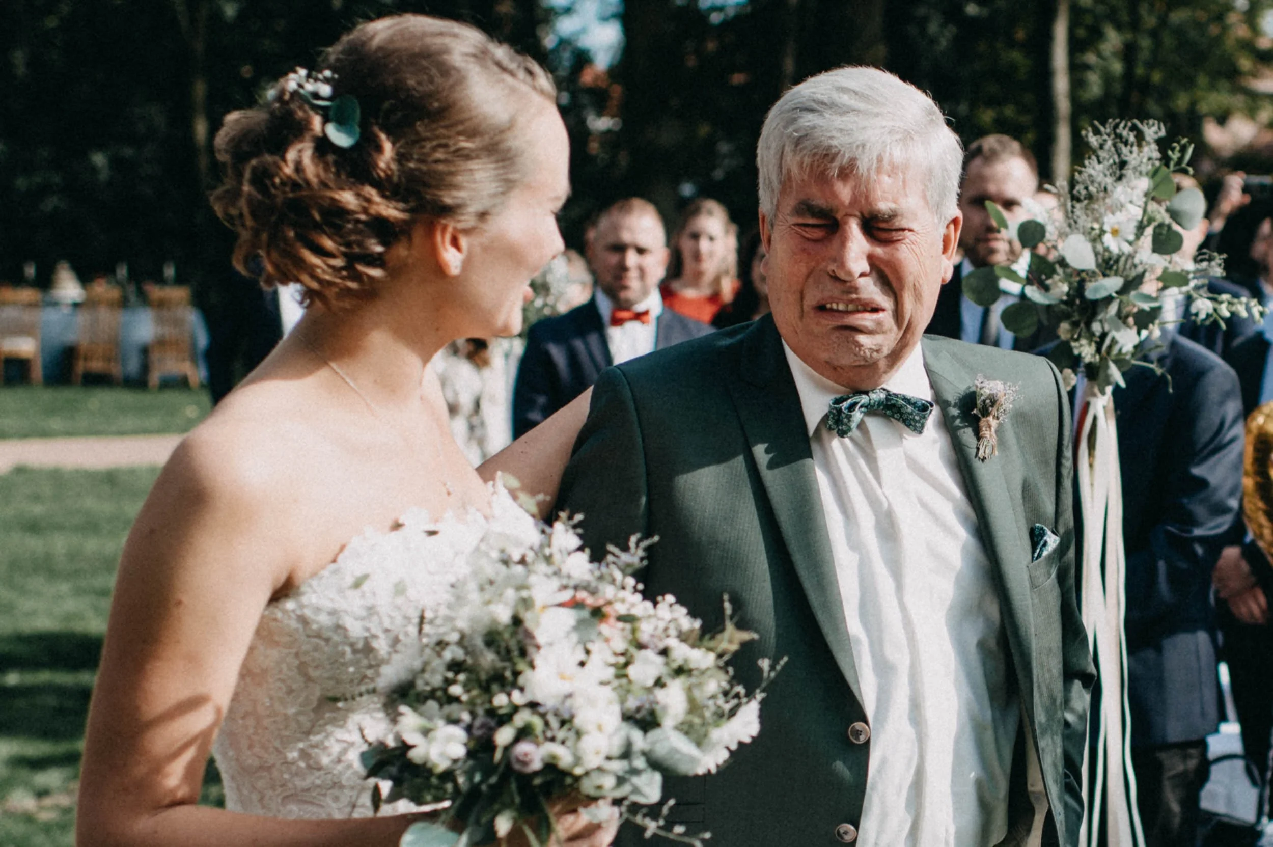 Ein Brautpaar bei einer Hochzeit im Freien, die Braut trägt ein weißes Spitzenkleid und hält einen Blumenstrauß, der Bräutigam trägt einen Anzug mit Fliege. Im Hintergrund sind Gäste sichtbar.