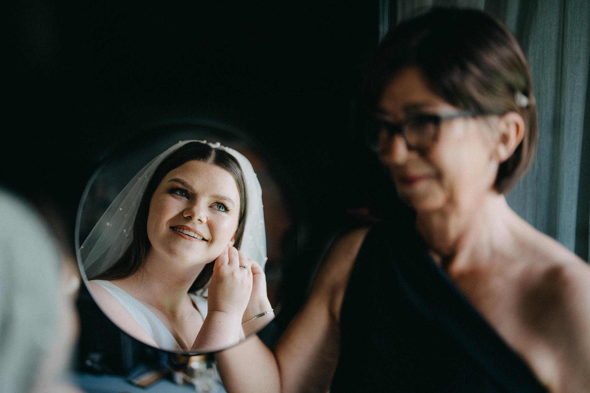 Braut betrachtet sich in einem Handspiegel, während eine andere Frau neben ihr lächelt.