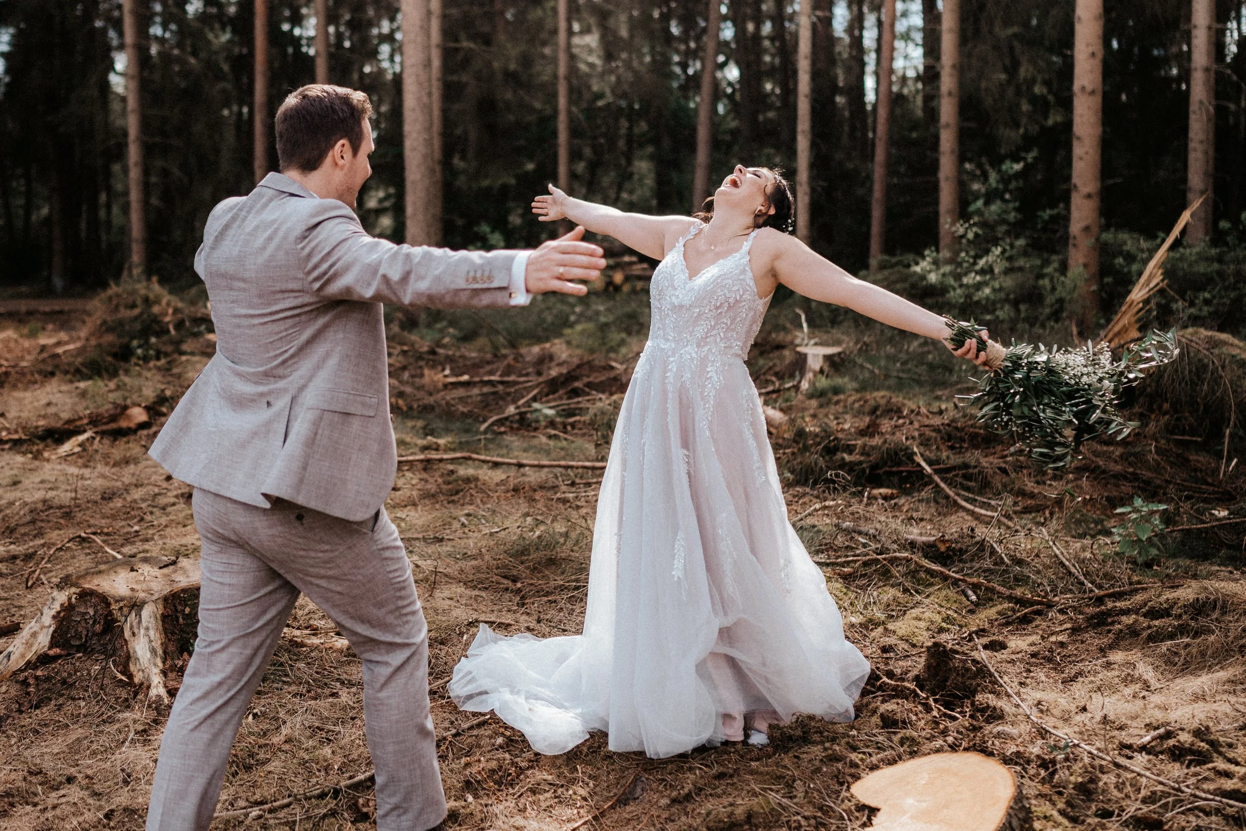 Ein Mann in einem hellgrauen Anzug und eine Braut in einem weißen Hochzeitskleid tanzen in einem Wald, sie lachen und haben die Arme weit ausgebreitet.