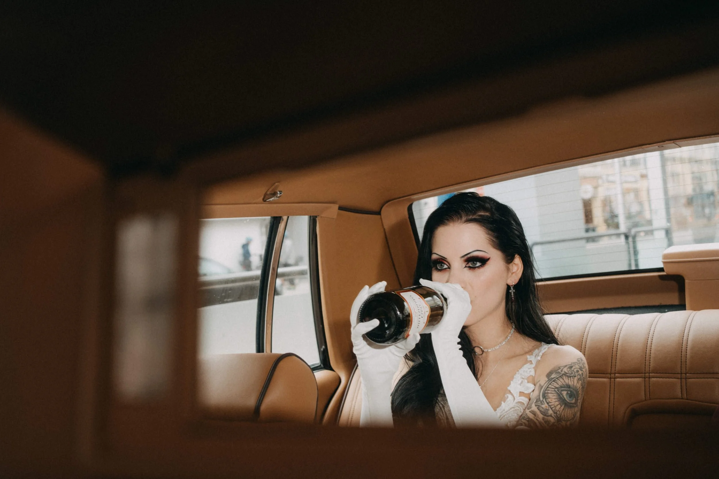 Eine Frau mit dunklen Haaren, Tattoo am Arm, trägt weiße Handschuhe und ein weißes Kleid, trinkt aus einer Flasche in einem alten Auto.