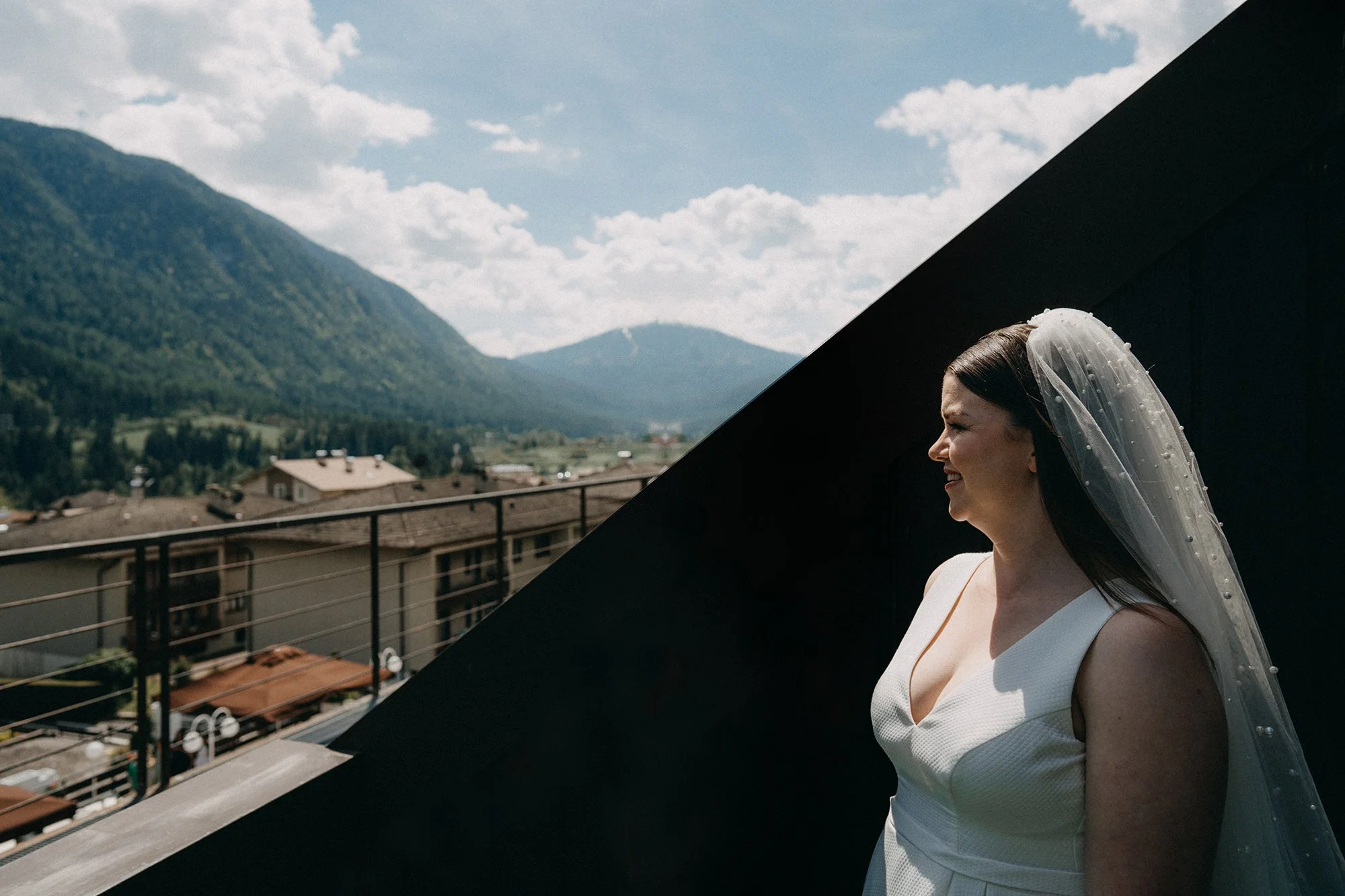 Eine Frau in Hochzeitskleidung mit Schleier steht auf einem Balkon mit Berglandschaft im Hintergrund. Sie lächelt und blickt nach rechts.