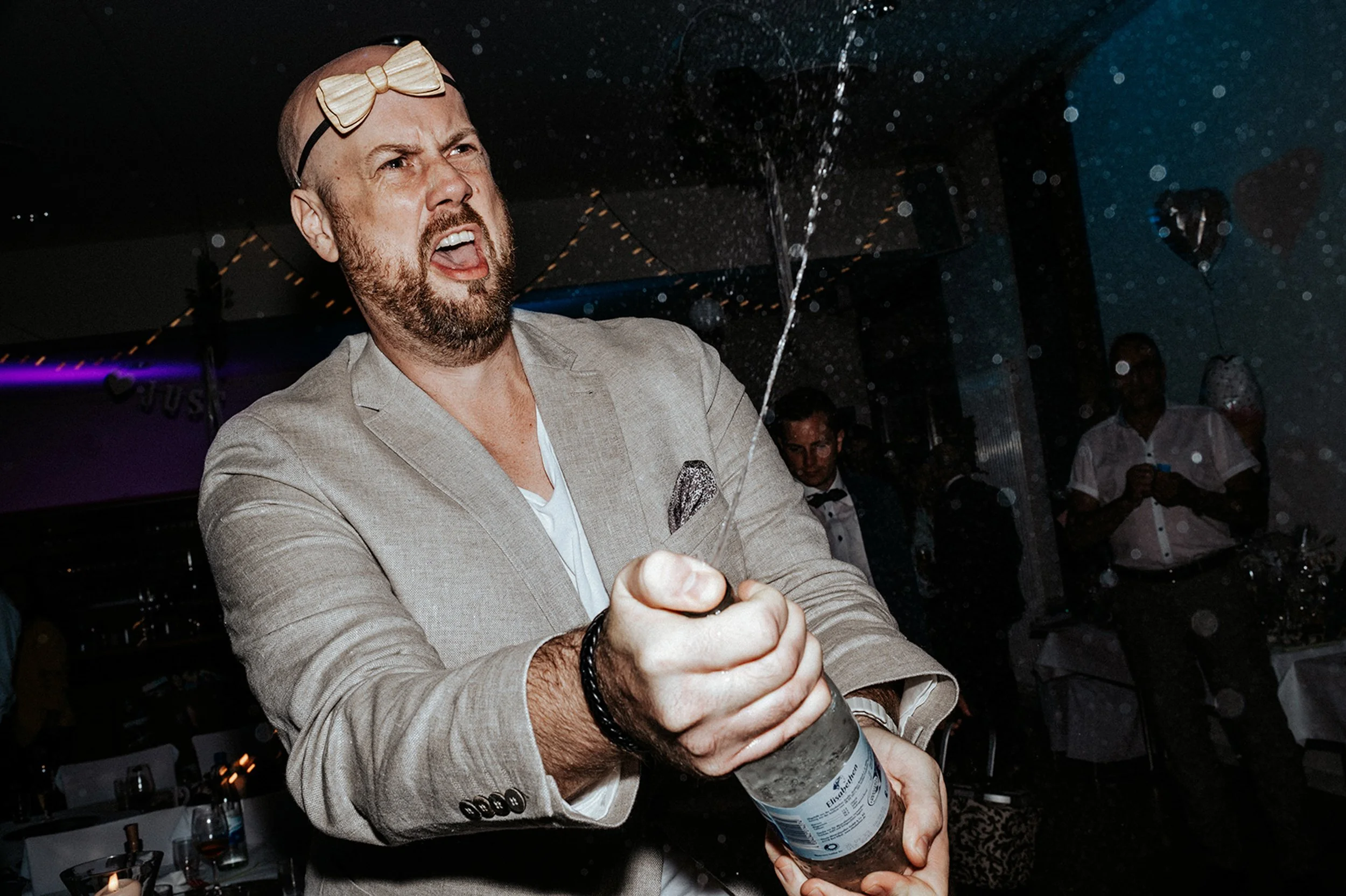 Ein Mann in einem Anzug öffnet mit einem Flaschenöffner eine Flasche, während eine Person im Hintergrund zuschaut.