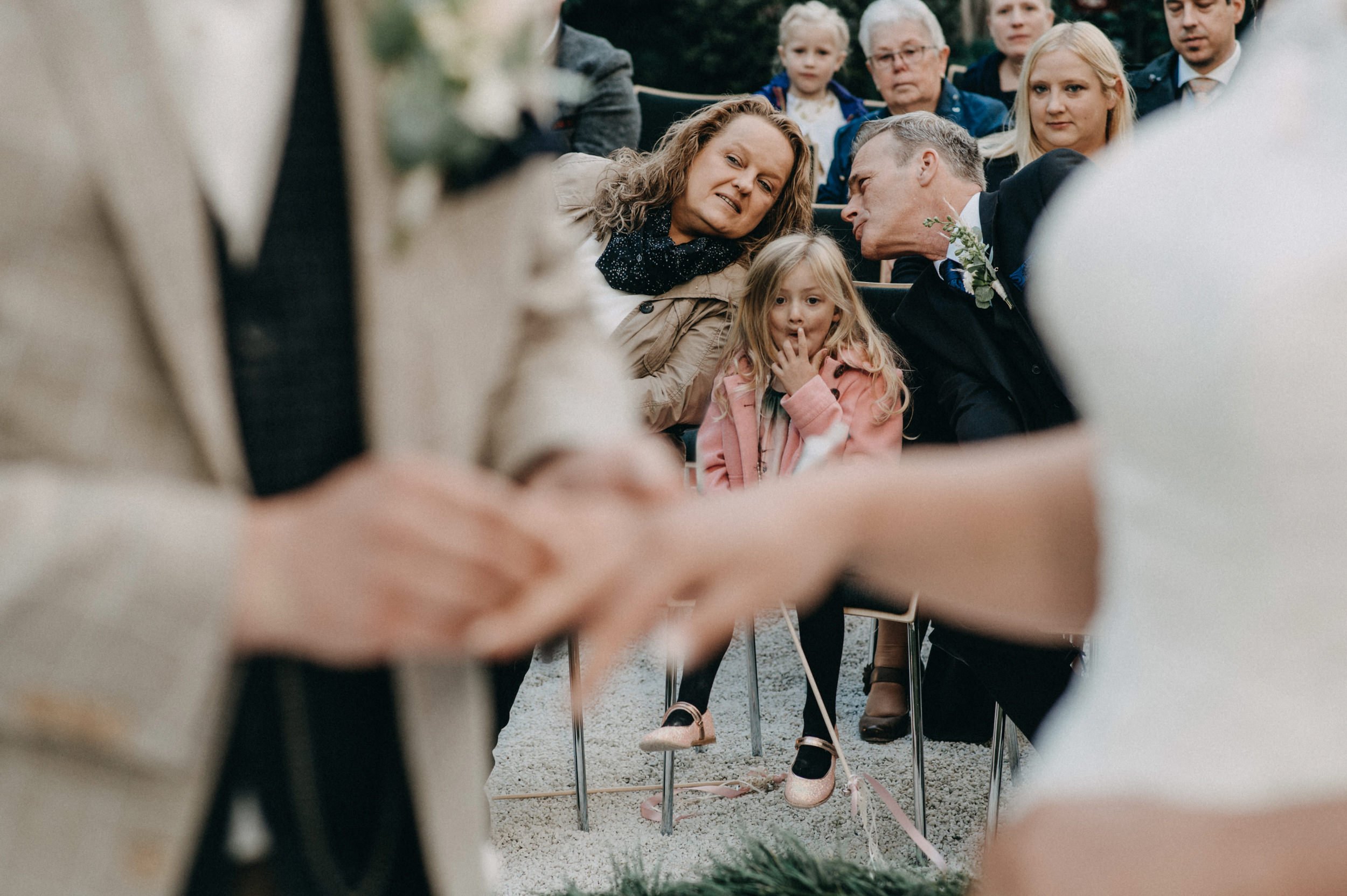 Menschen sitzen bei einer Zeremonie im Freien, im Mittelpunkt ein Mädchen in rosa Jacke und Schuhe, umgeben von Erwachsenen, einige schauen ernst.