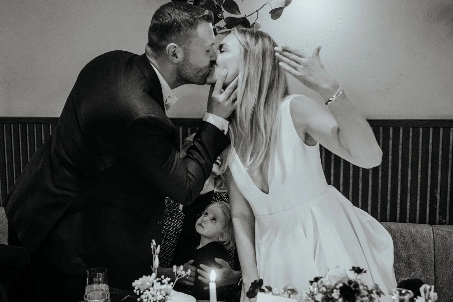 Ein Mann und eine Frau küssen sich bei einer Feier, während ein kleines Kind sie anschaut. Es gibt einen Tisch mit Blumen und einer brennenden Kerze im Vordergrund.