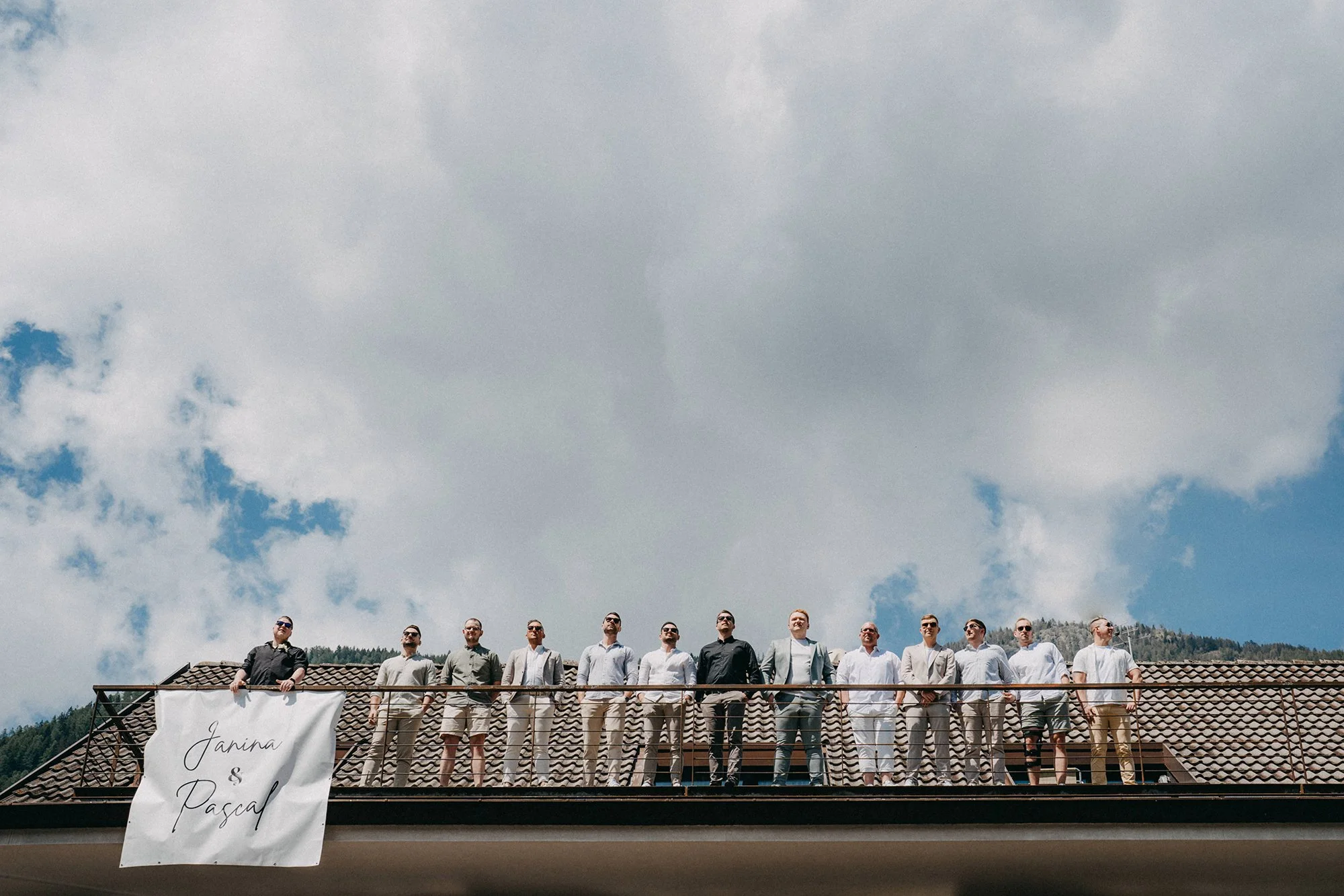 Eine Gruppe von Männern steht auf einem Balkon unter einem klaren Himmel mit Wolken. Es scheint eine formelle Veranstaltung oder ein Anlass zu sein, wahrscheinlich eine Hochzeit, da ein Banner mit den Namen "Janina & Pascal" sichtbar ist.