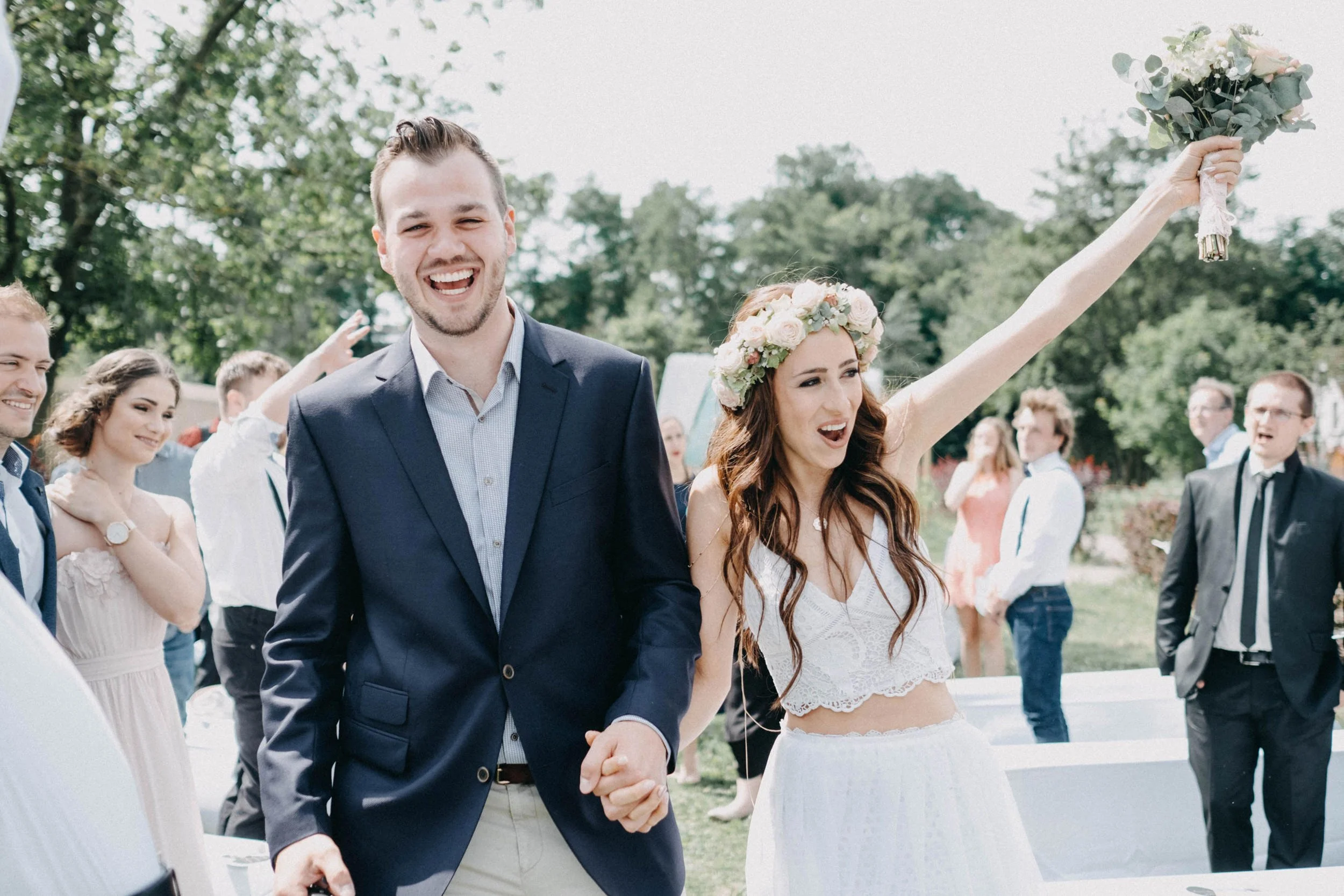 Ein lachendes Brautpaar bei einer Hochzeit im Freien, umgeben von Gästen, die sich freuen. Die Braut trägt einen Blumenkranz und ein Spitzenkleid, der Bräutigam trägt einen Anzug.