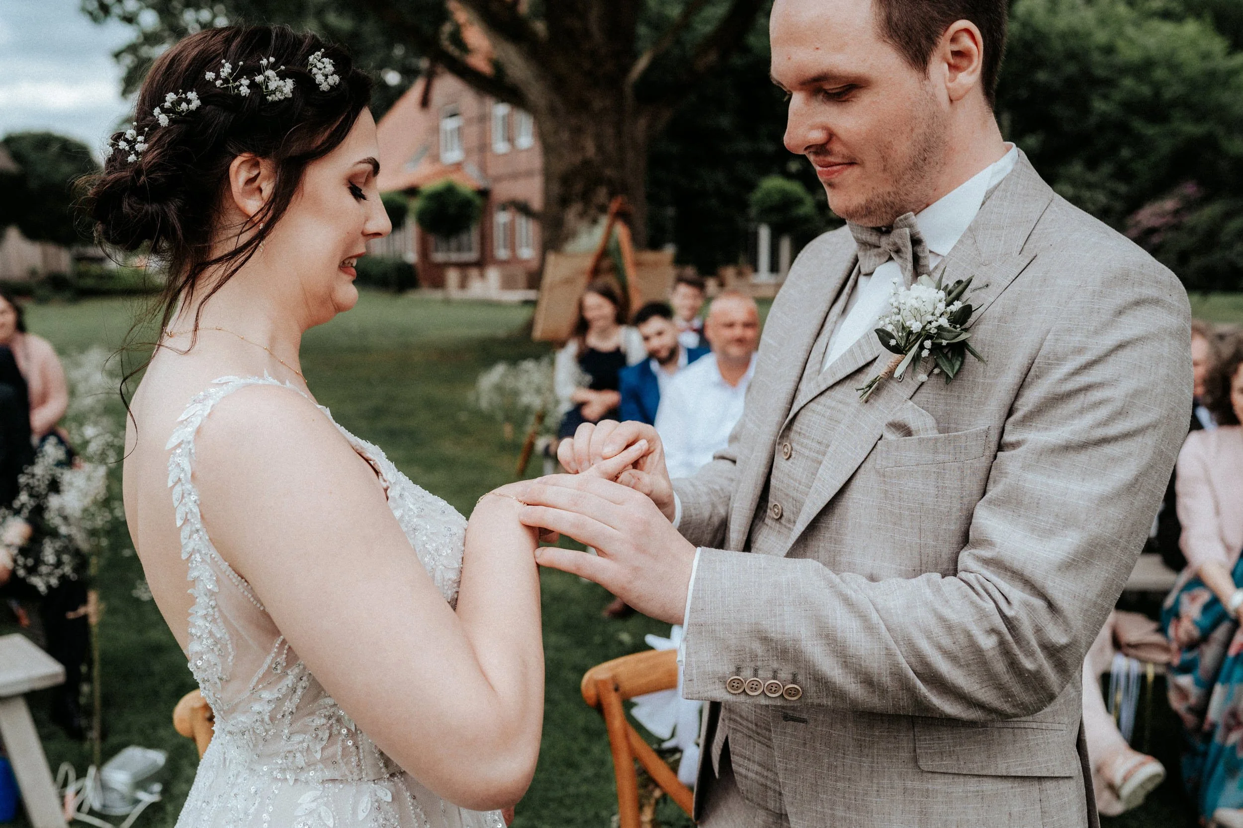 Ein Brautpaar bei einer Hochzeit im Freien: Die Braut trägt ein weißes Hochzeitskleid mit Spitze, die Figur des Bräutigams ist in einem hellgrauen Anzug mit Fliege. Sie halten Händchen und blicken liebevoll aufeinander, im Hintergrund sind Blumen, Bä