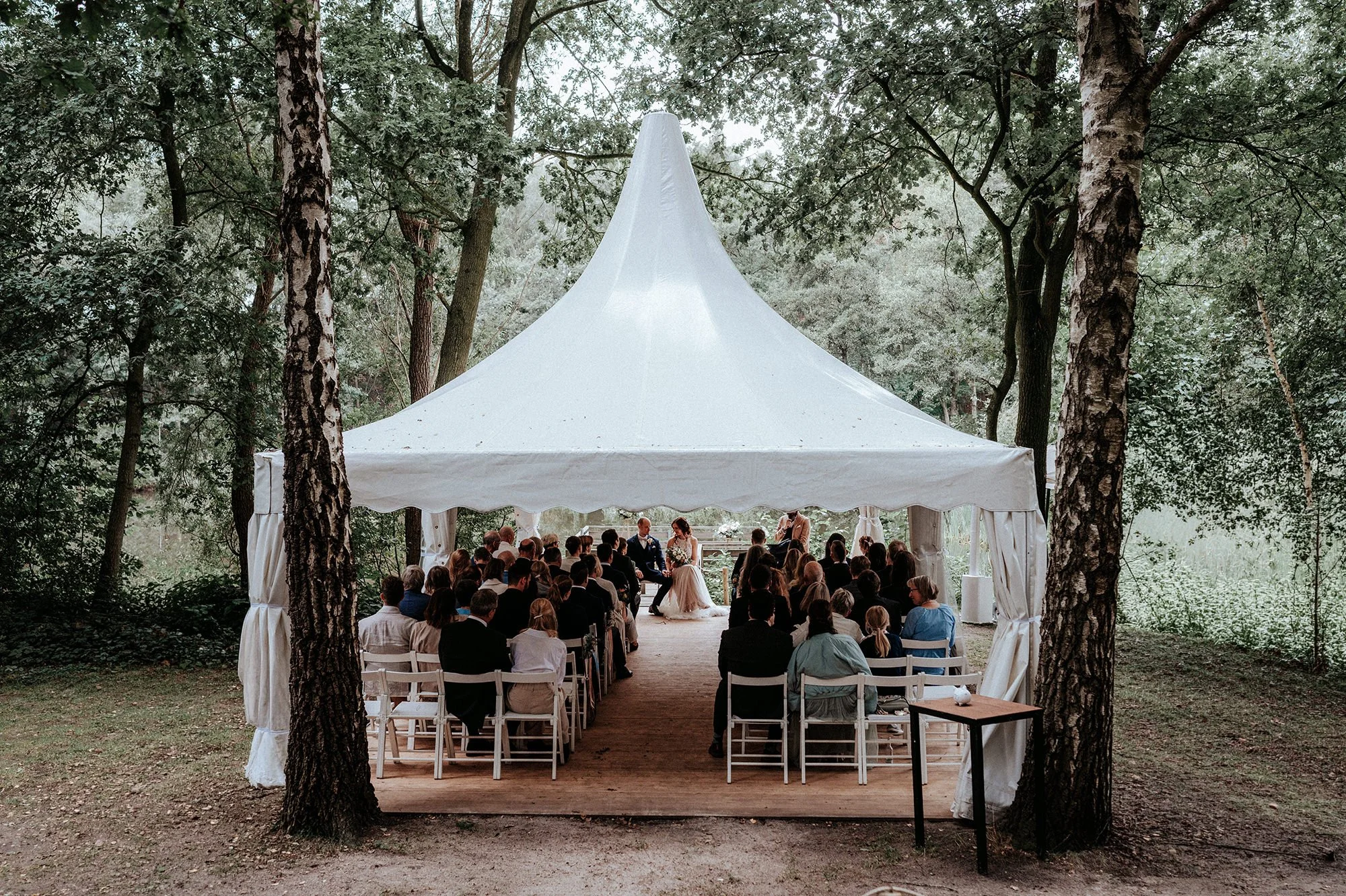 Hochzeit unter einem weißen Zelt inmitten eines Waldes mit vielen Gästen, einem Brautpaar und Trauzeugen