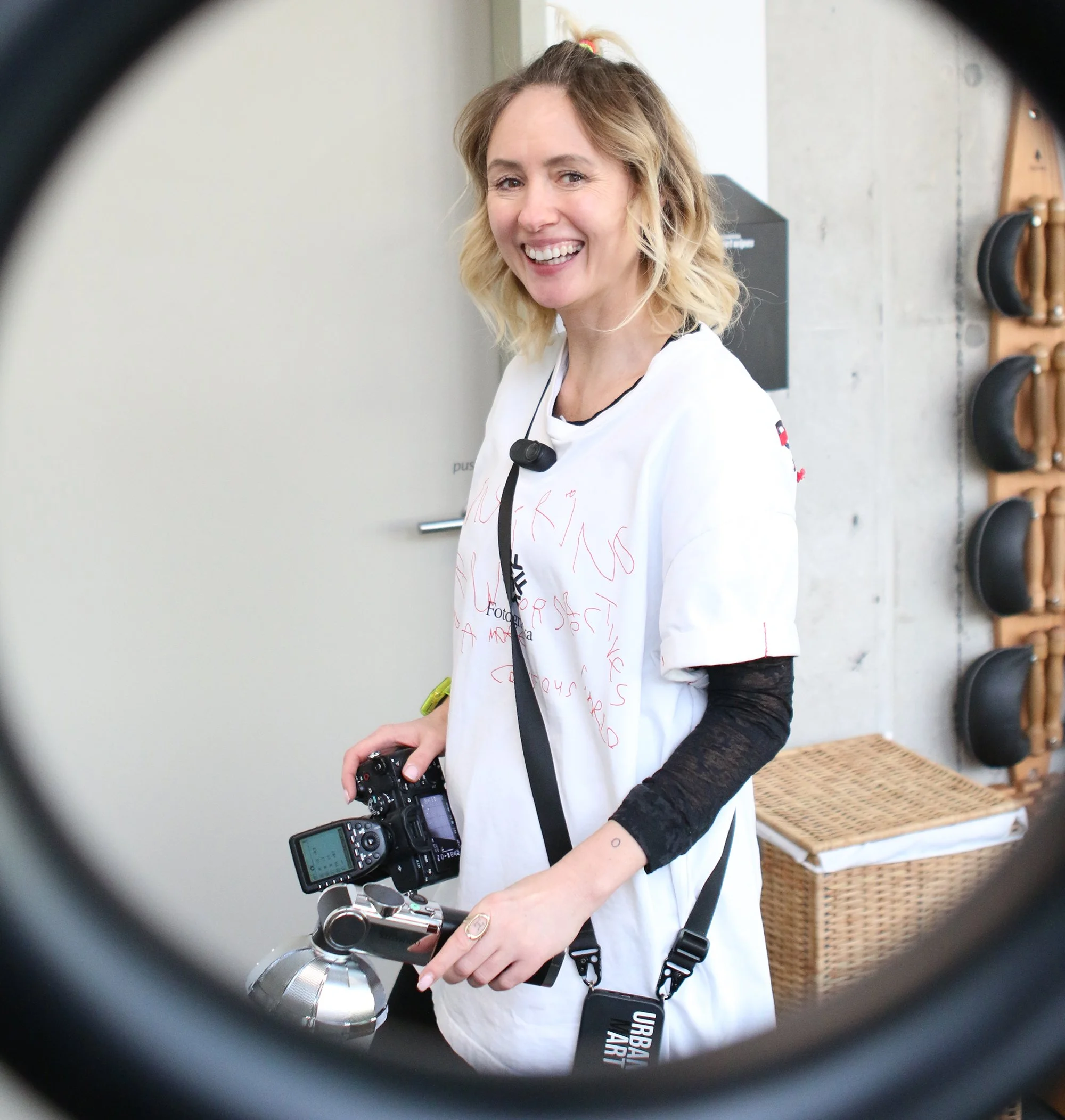 Eine lächelnde Frau wird durch ein Fernglas fotografiert, umgeben von Kameraausrüstung, im Hintergrund eine Wand mit Wandhaltern für Schuhe.