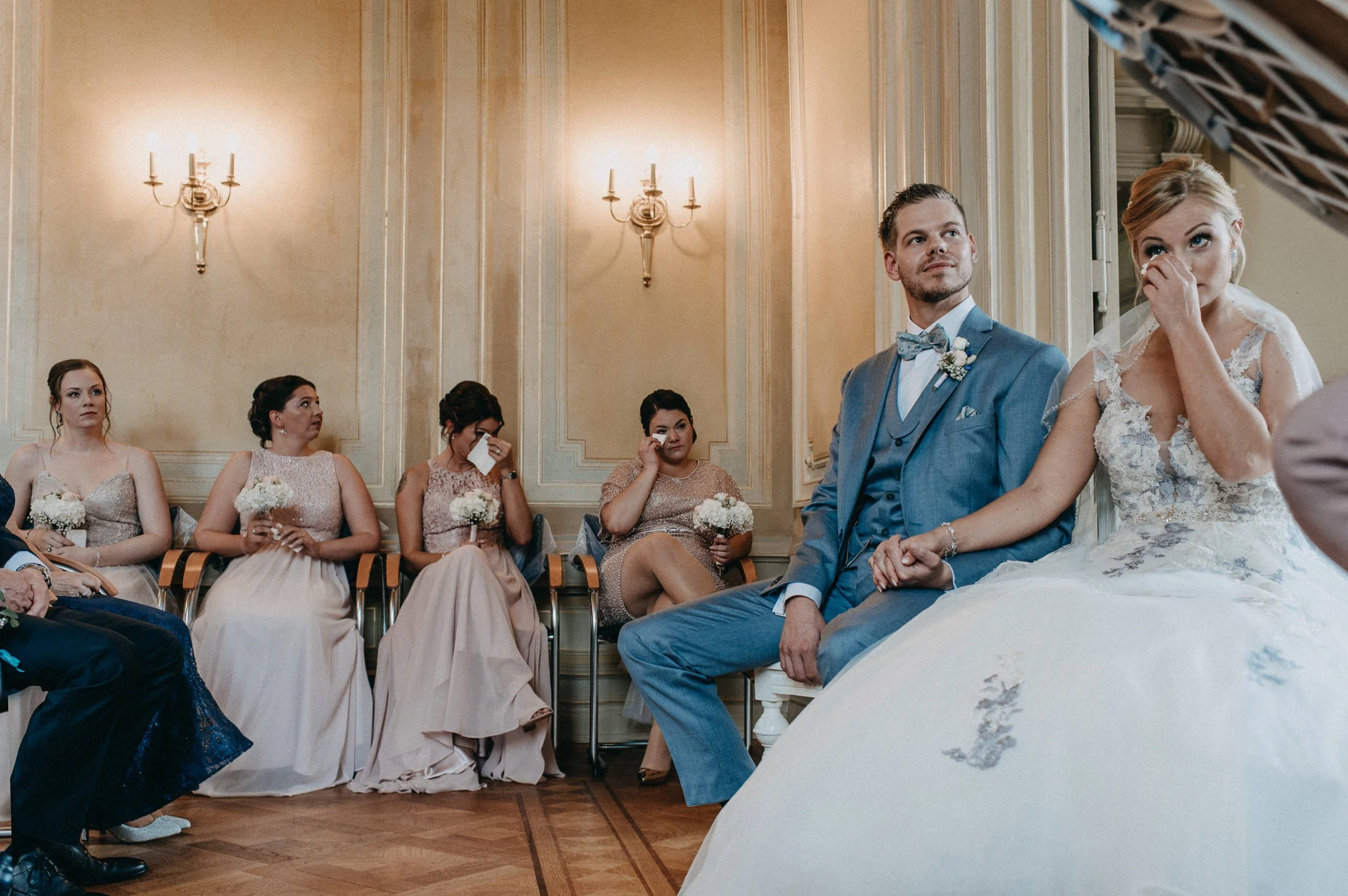 Hochzeitsszene in einem eleganten Saal, Braut und Bräutigam sitzen in der ersten Reihe, umgeben von ihren Hochzeitsgästen, die emotionale Momente erleben.