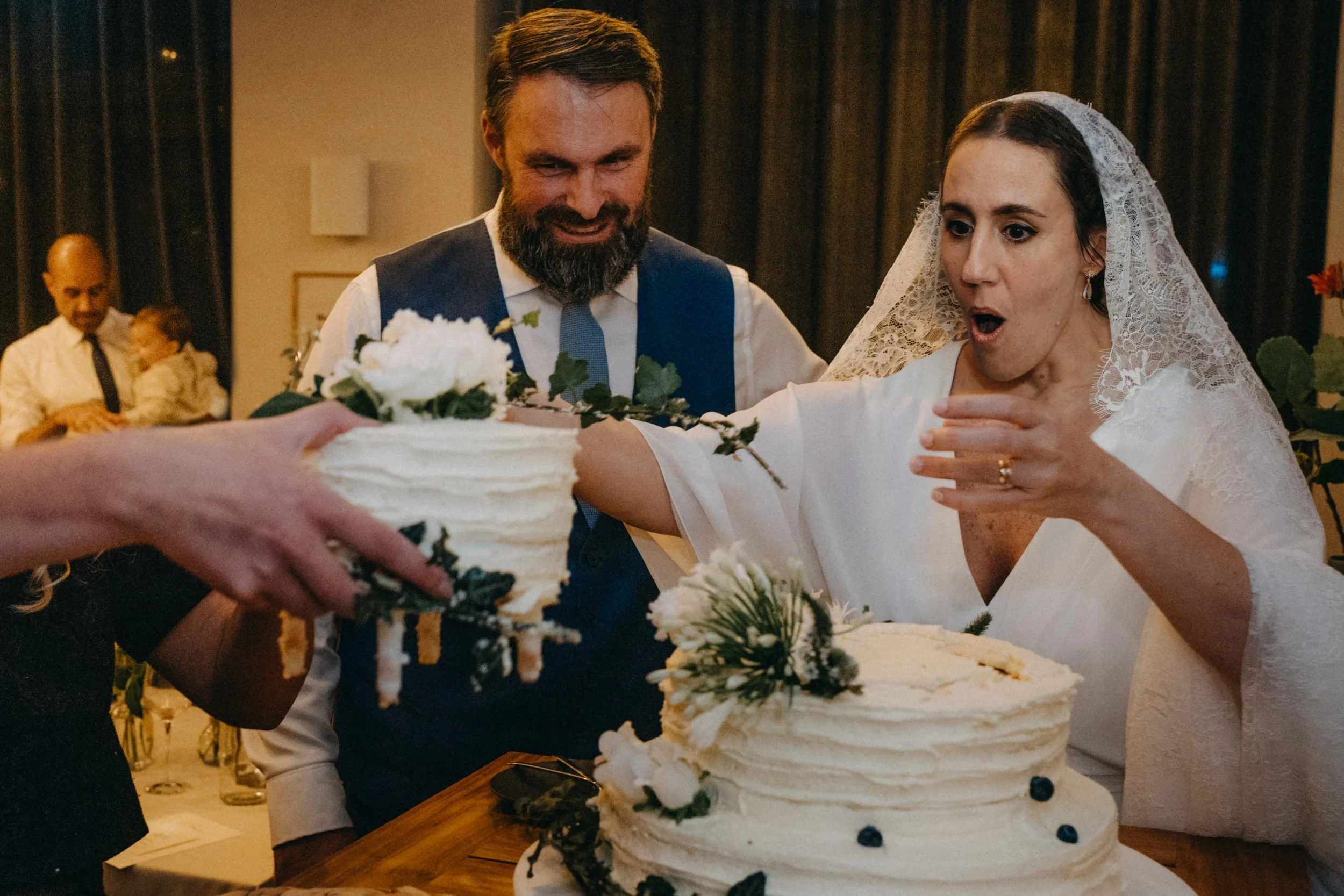 Ein Brautpaar beim Anschneiden der Hochzeitstorte, die mit weißen Blumen und grünem Laub dekoriert ist.