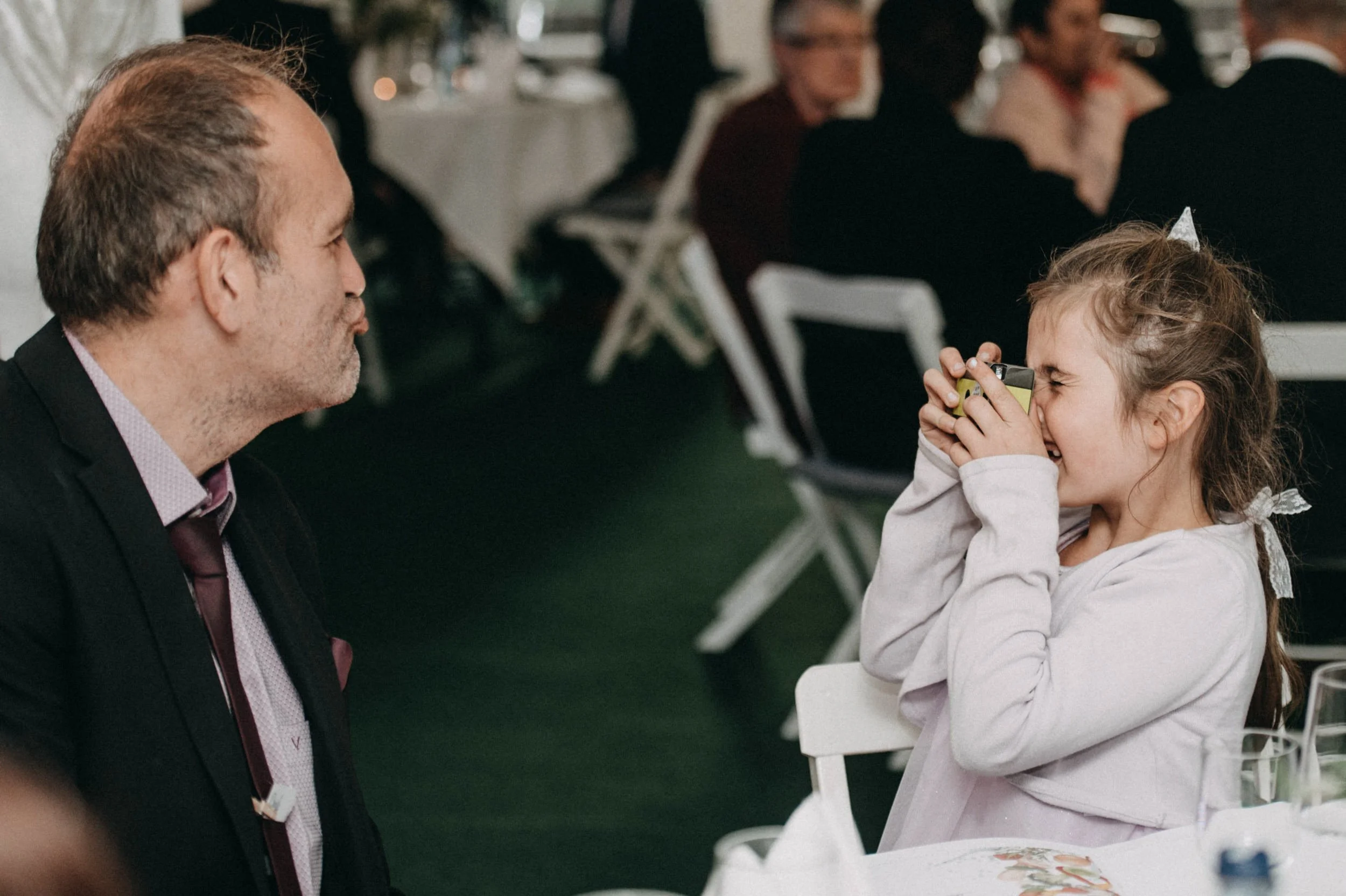 Ein Mann im Anzug lächelt einer lachenden kleinen Mädchen, die Mädchen trägt eine weiße Jacke und hält eine Tasse vor das Gesicht; im Hintergrund sitzen andere Menschen bei einem Essen in einem festlichen Rahmen.