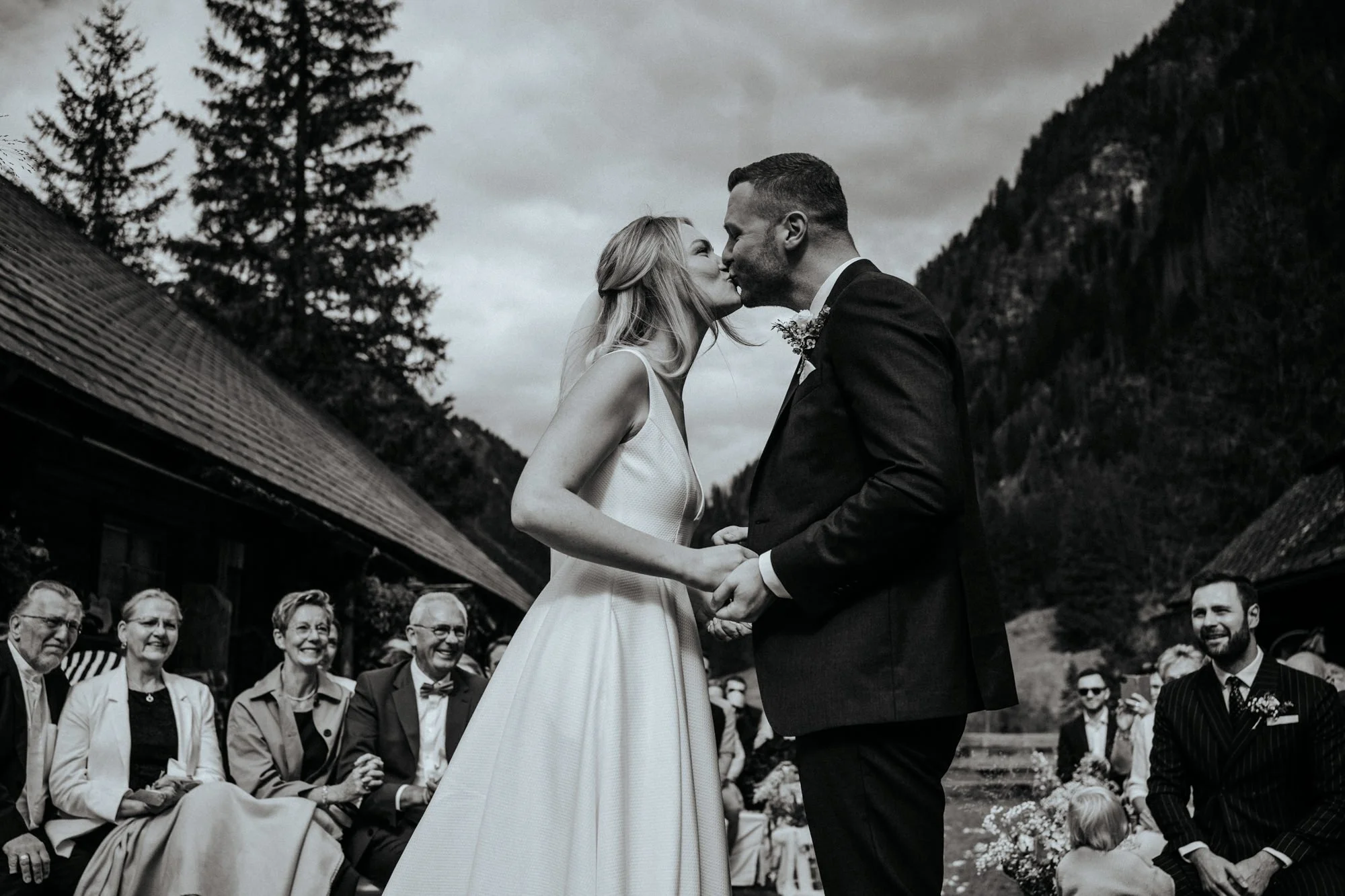 Ein Brautpaar bei einer Hochzeit, es küsst sich, während eine Gruppe von Gästen im Hintergrund zusieht. Die Szene ist in einer naturnahen Umgebung mit Bergen und Bäumen.