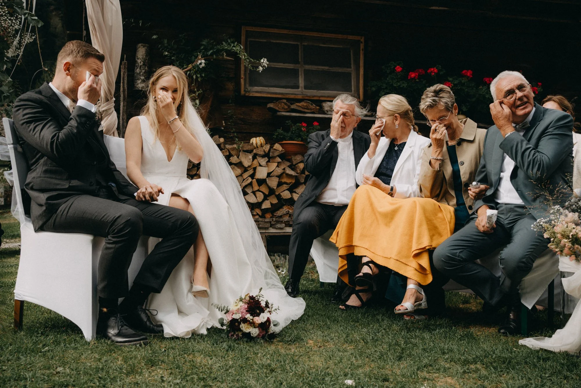Junge Braut und Bräutigam sitzen bei einer emotionalen Trauungszeremonie, umgeben von Freunden und Familie, in einem rustikalen Außenbereich mit Holz und Blumen.