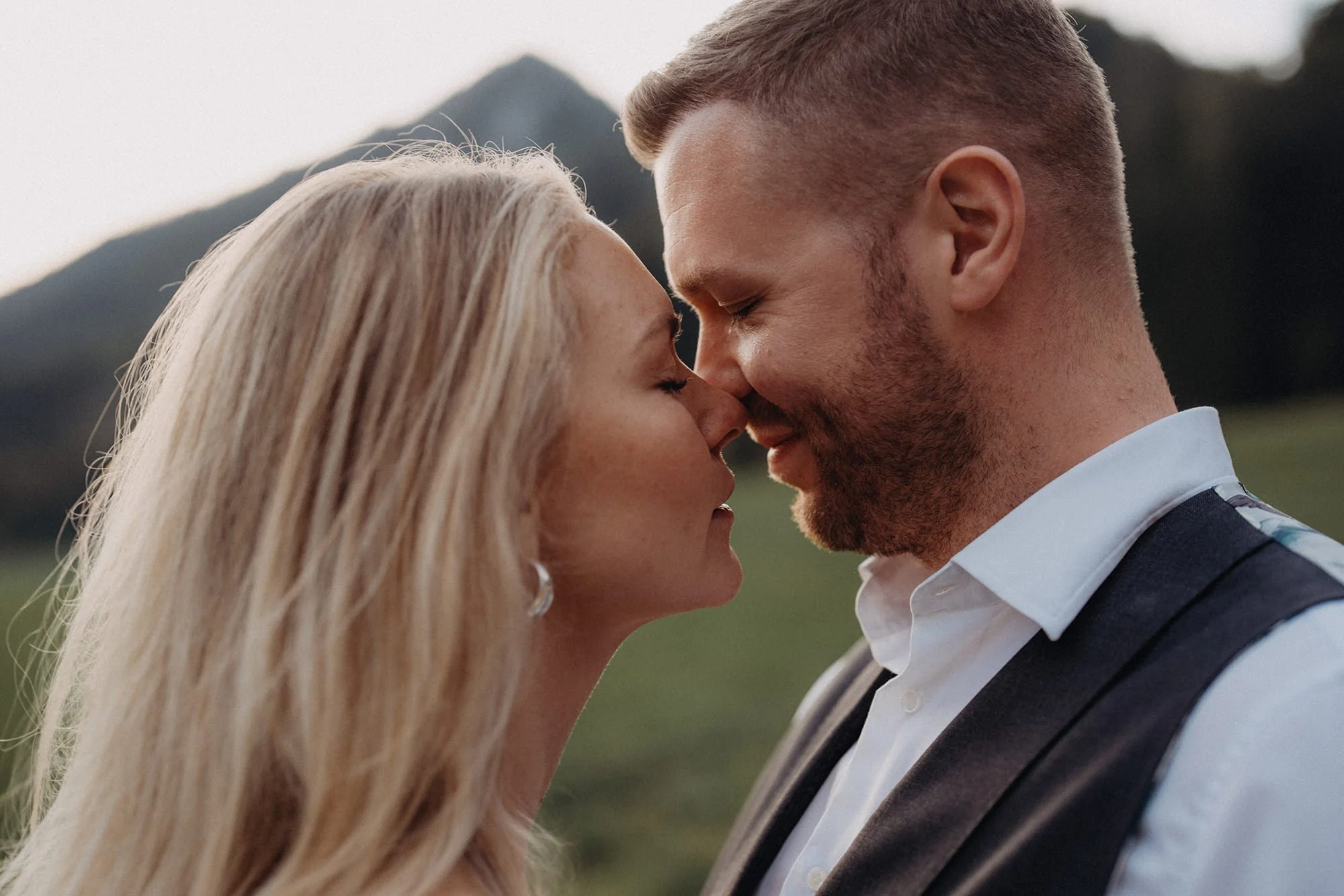 Ein verliebtes Paar steht sich mit geschlossen Augen und Stirn an Stirn im Freien. Der Mann trägt einen weißen Hemd mit dunklem Sakko und hat kurzes Haar. Das Mädchen hat lange blonde Haare und trägt ein helles Kleid.