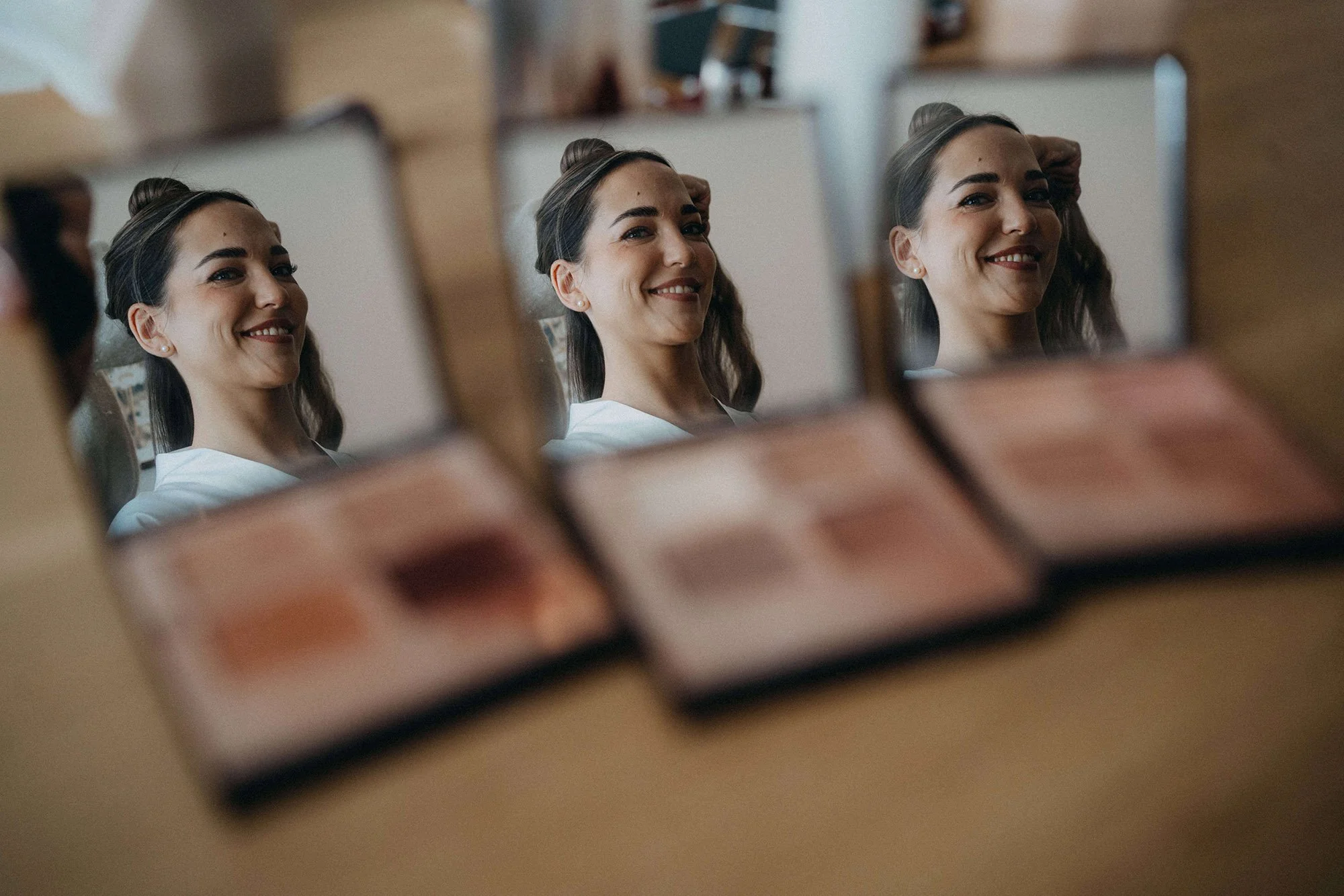 Nahaufnahme eines Make-up-Paletten mit verschiedenen Lidschattenfarben, im Vordergrund unscharf, im Hintergrund eine lächelnde Frau, die auf einen Spiegel blickt.