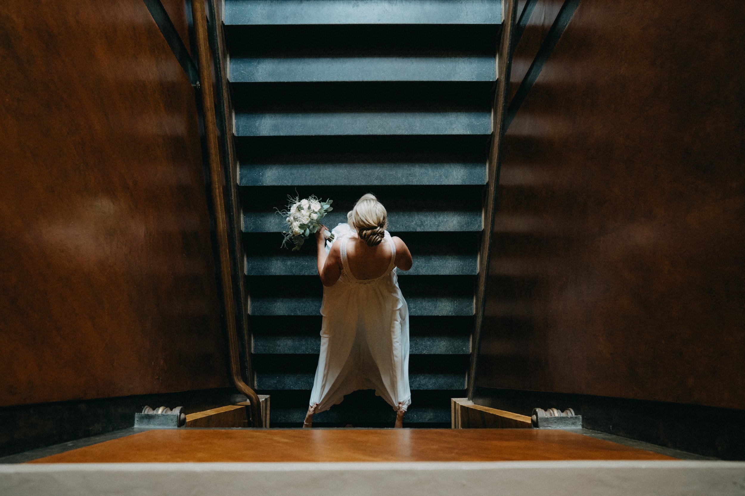 Eine Braut in einem weißen Kleid steht auf einer Treppe, die von dunklen Holzwänden flankiert wird, und hält einen Blumenstrauß in der Hand.