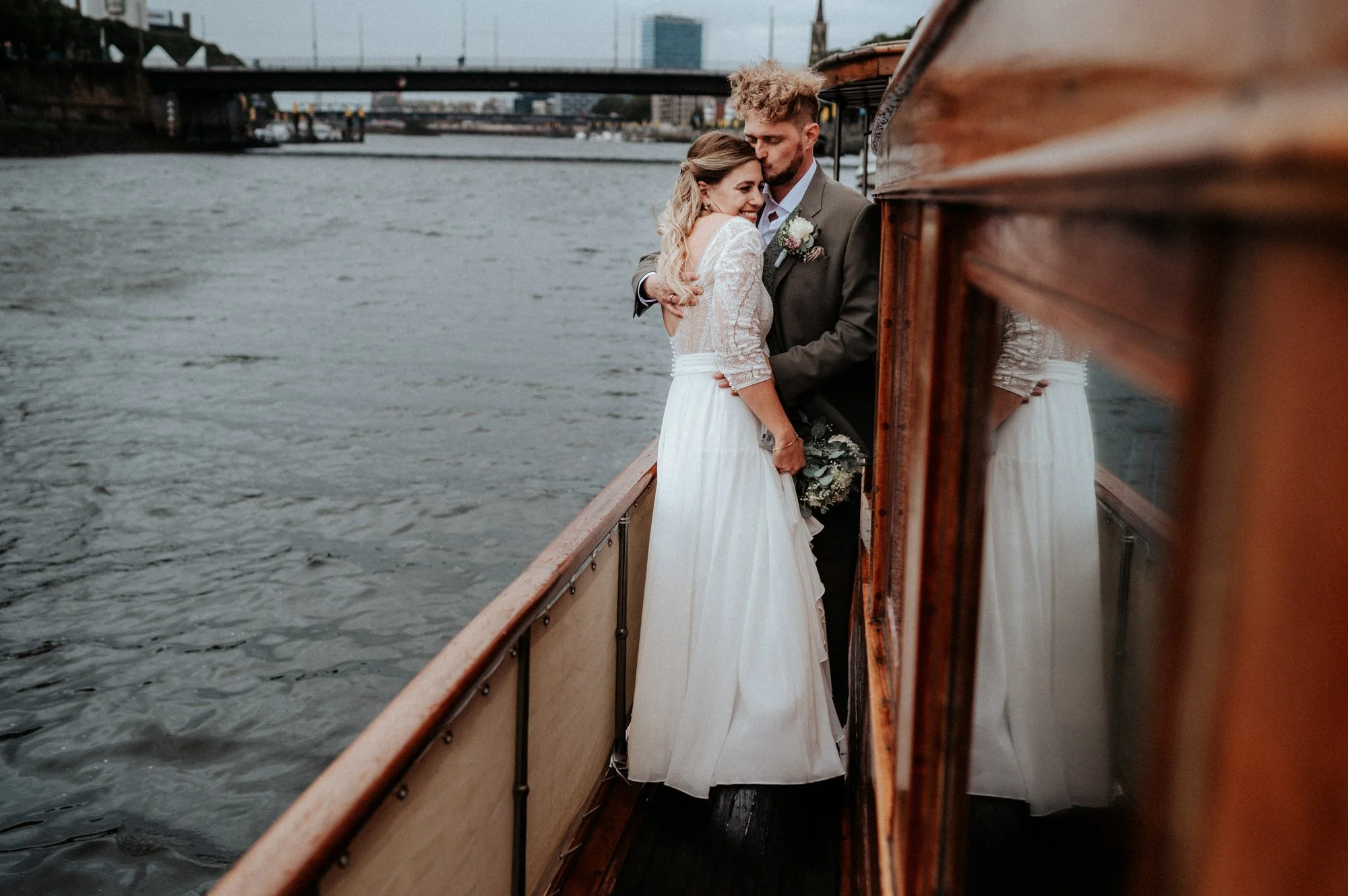 Ein Paar bei einer Hochzeit auf einem Boot auf einem Fluss, umgeben von einer städtischen Landschaft mit Brücken und Hochhäusern, bei bewölktem Himmel.
