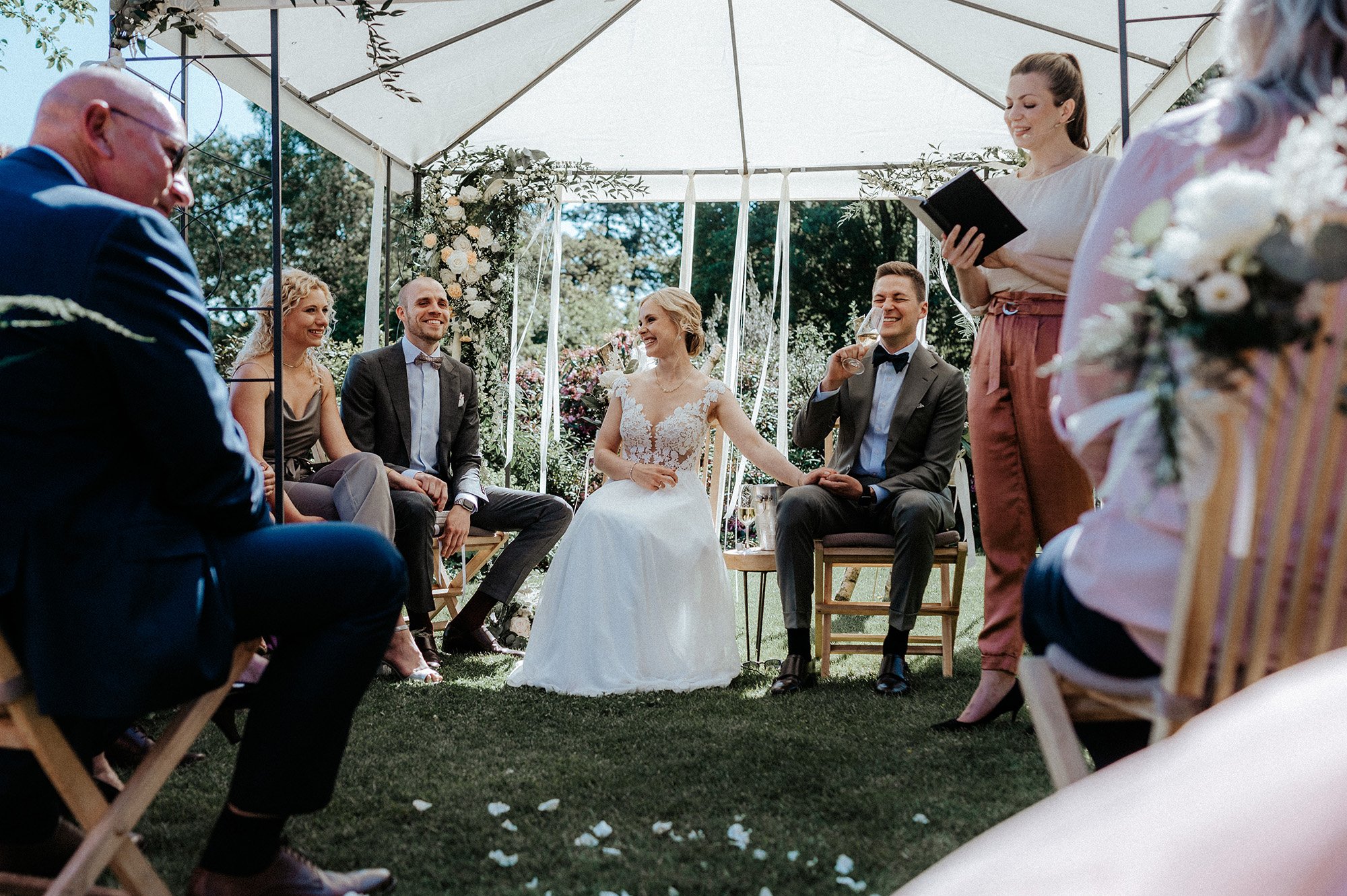 Hochzeit im Garten mit six Personen unter einem weißen Zelt, Blüten und grüne Bäume im Hintergrund, Braut, Bräutigam und Gäste, die Zeugen und Unterstützung bei der Zeremonie sind.