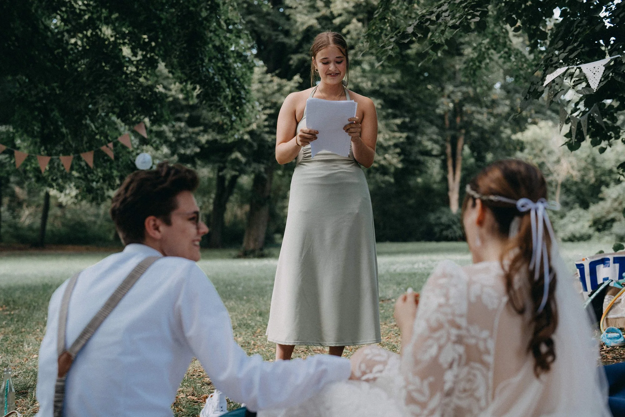 Junge Frau liest an einer Outdoor-Feier im Park, umgeben von Freunden, unter grünen Bäumen, mit dekorativen Girlanden im Hintergrund.