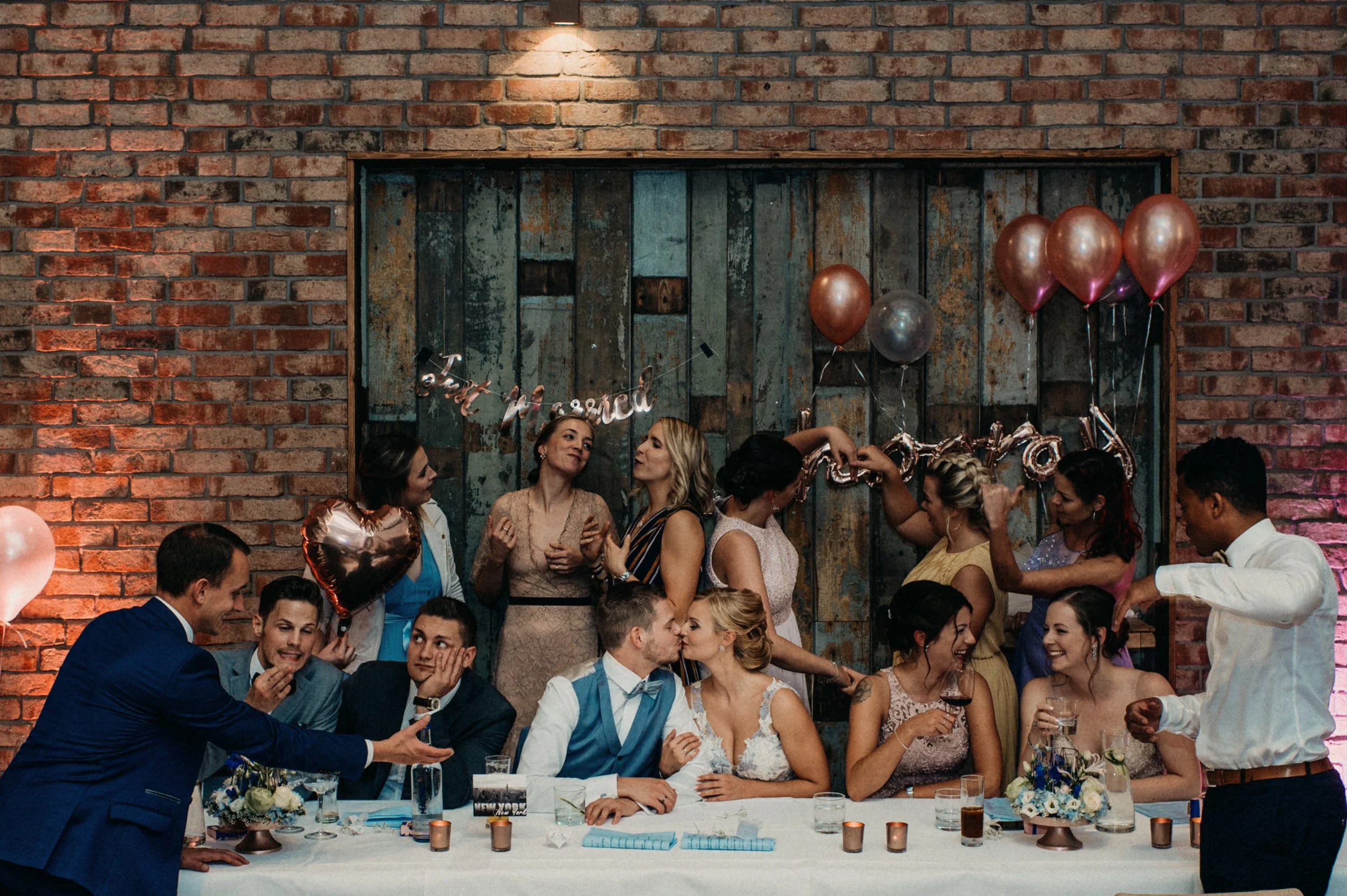 Feiernde Menschen bei einer Hochzeit oder Party, mit Ballons und Dekorationen vor einem Backsteinwand-Hintergrund.