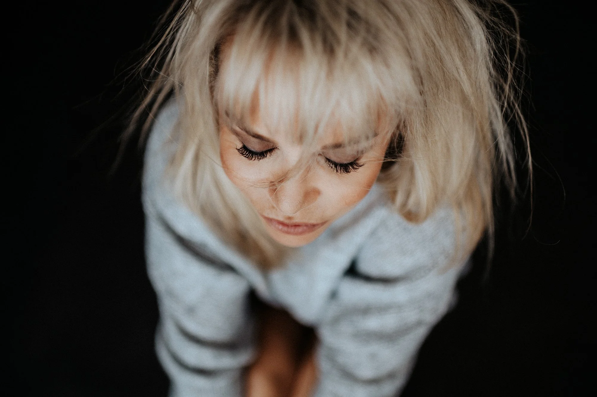 Nahaufnahme eines Mädchens mit geschlossenen Augen, blonde Haare, dunklem Hintergrund und einem weißen Pullover.