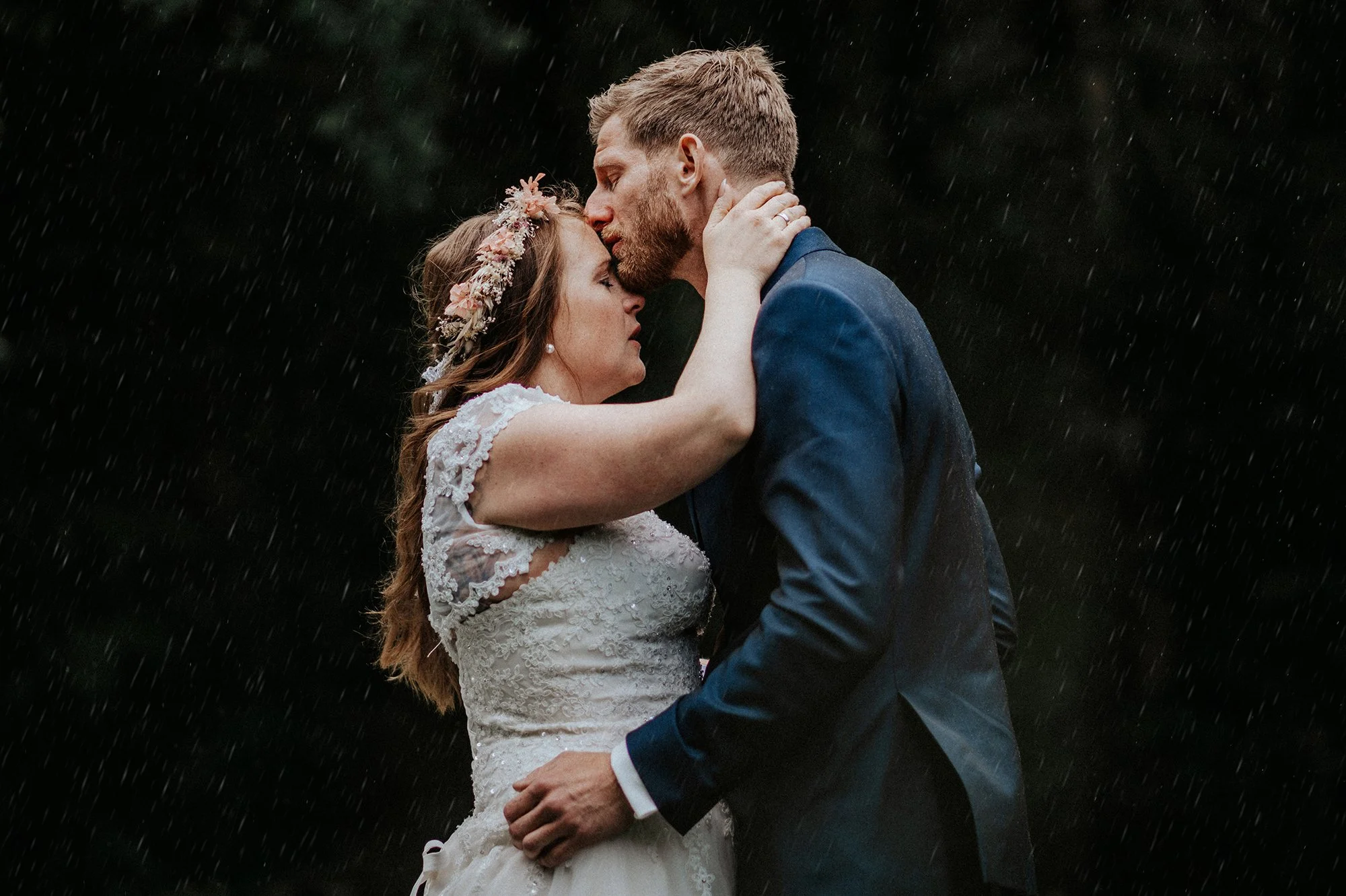 Ein Brautpaar bei einem Regen Schau spielt sich im Freien ab, sie küsst sich, er trägt einen blauen Anzug, sie trägt ein weißes Hochzeitskleid mit Spitzen, sie trägt eine Blumenkranz im Haar.