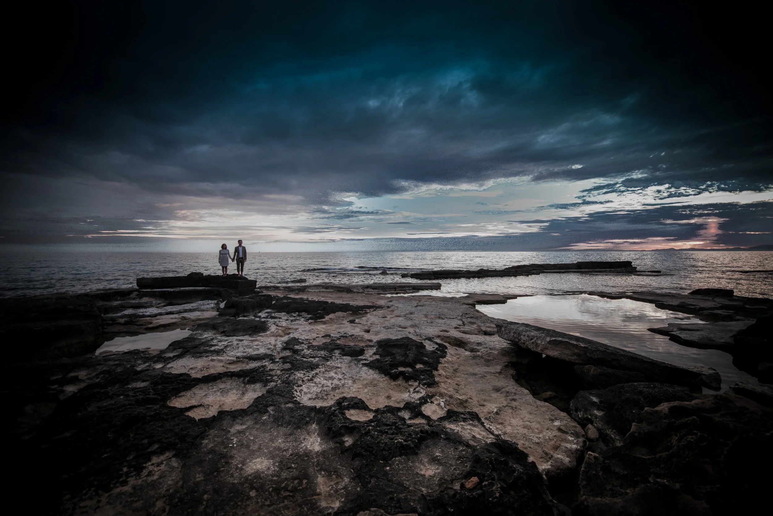 Zwei Menschen halten sich an den Händen und stehen auf Felsen am Meer bei Sonnenuntergang, dunkle Wolken am Himmel.